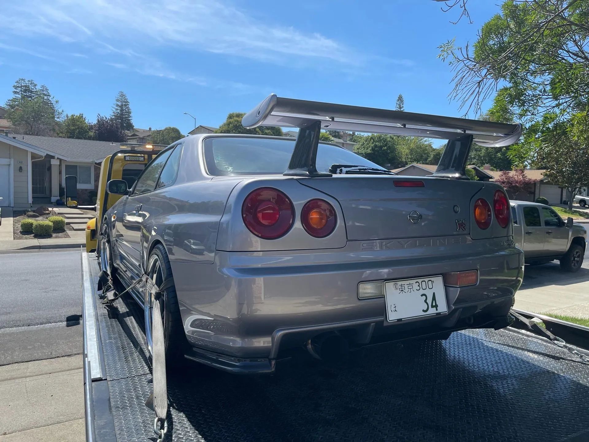 Grey Nissan Skyline R34 with large spoiler on a flatbed tow truck in a suburban area.