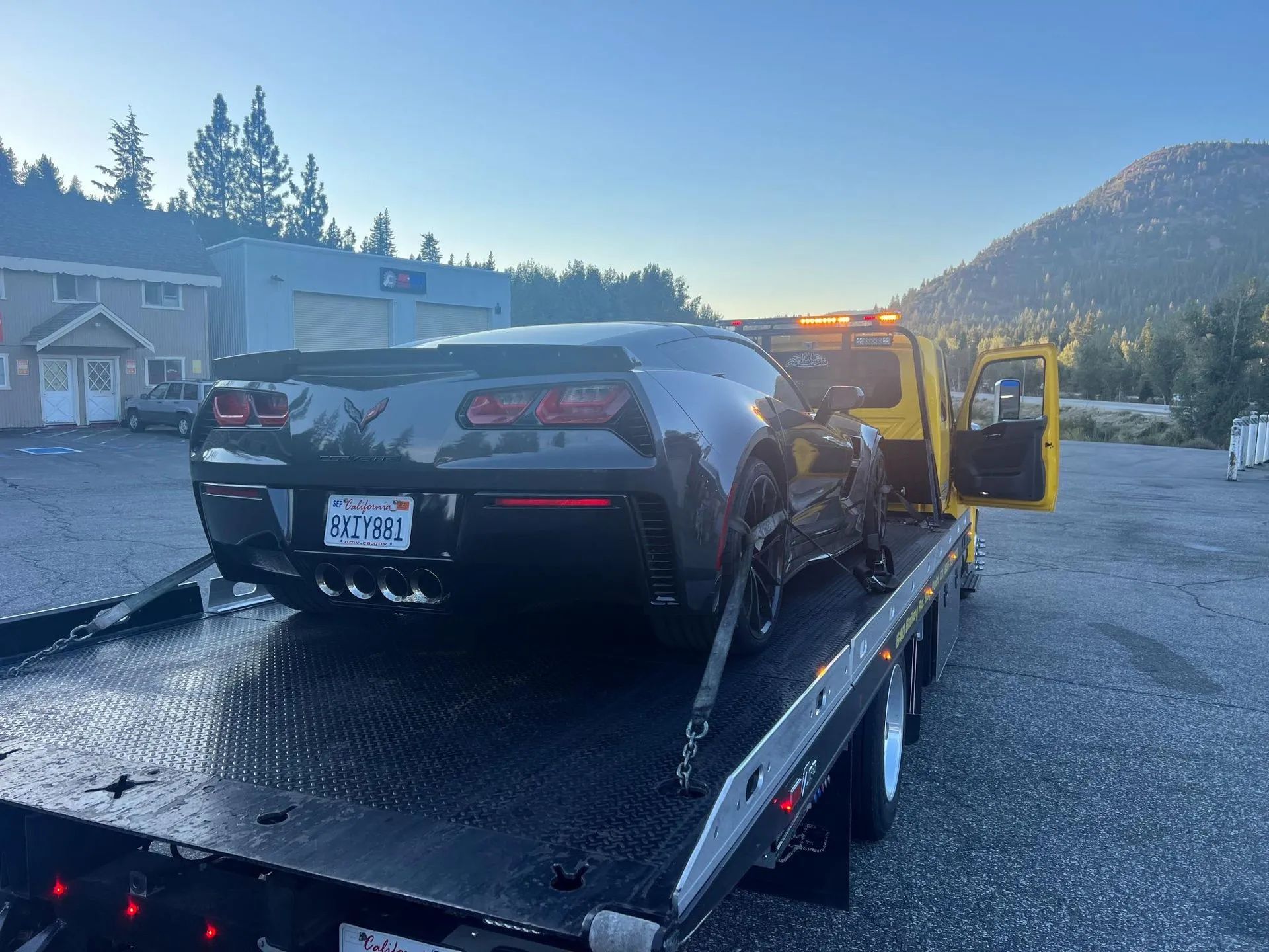 Gray sports car on a yellow tow truck bed, damaged front. Outdoors, building in background, sunny.