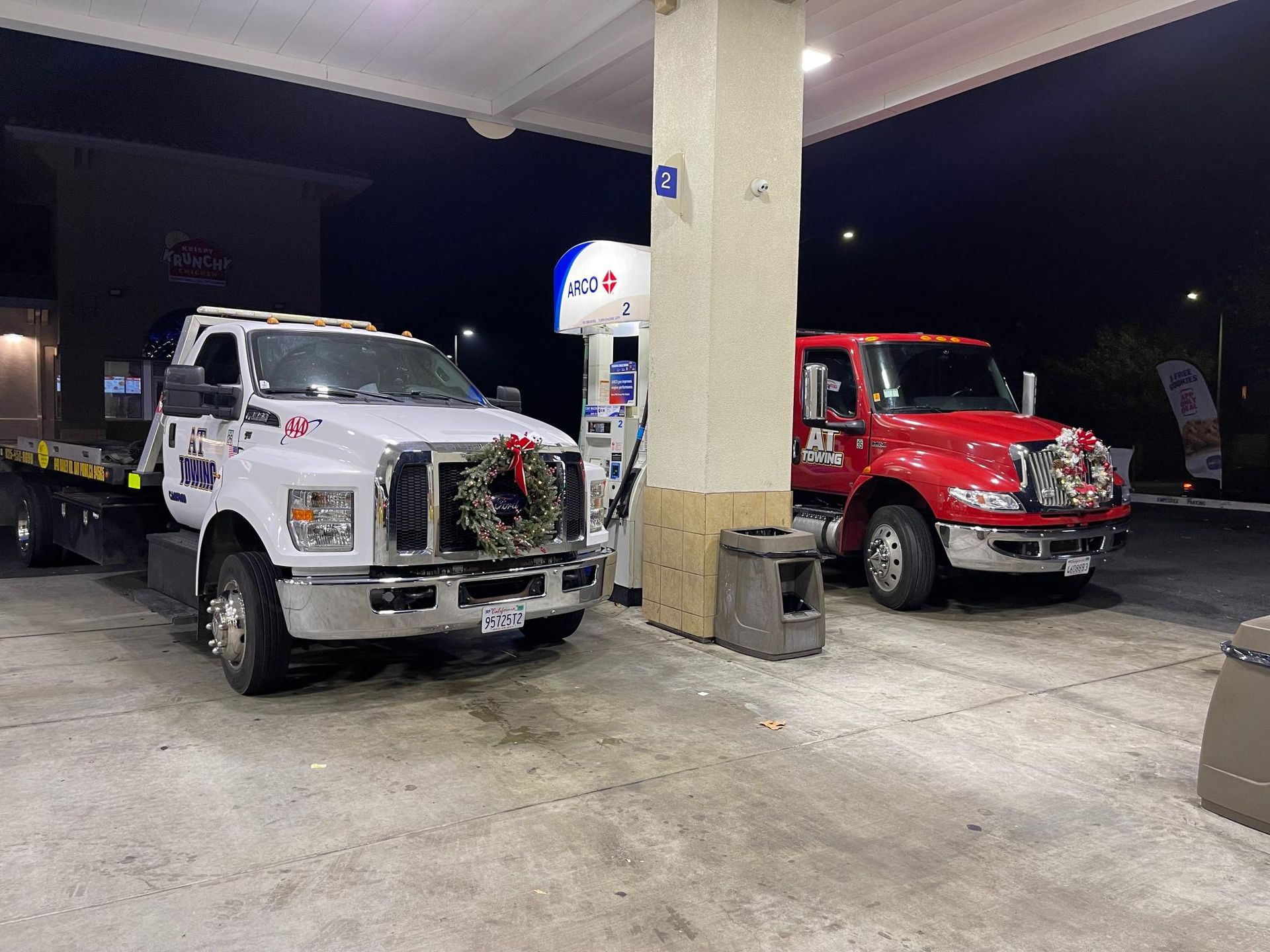 Two tow trucks are parked in a gas station at night.