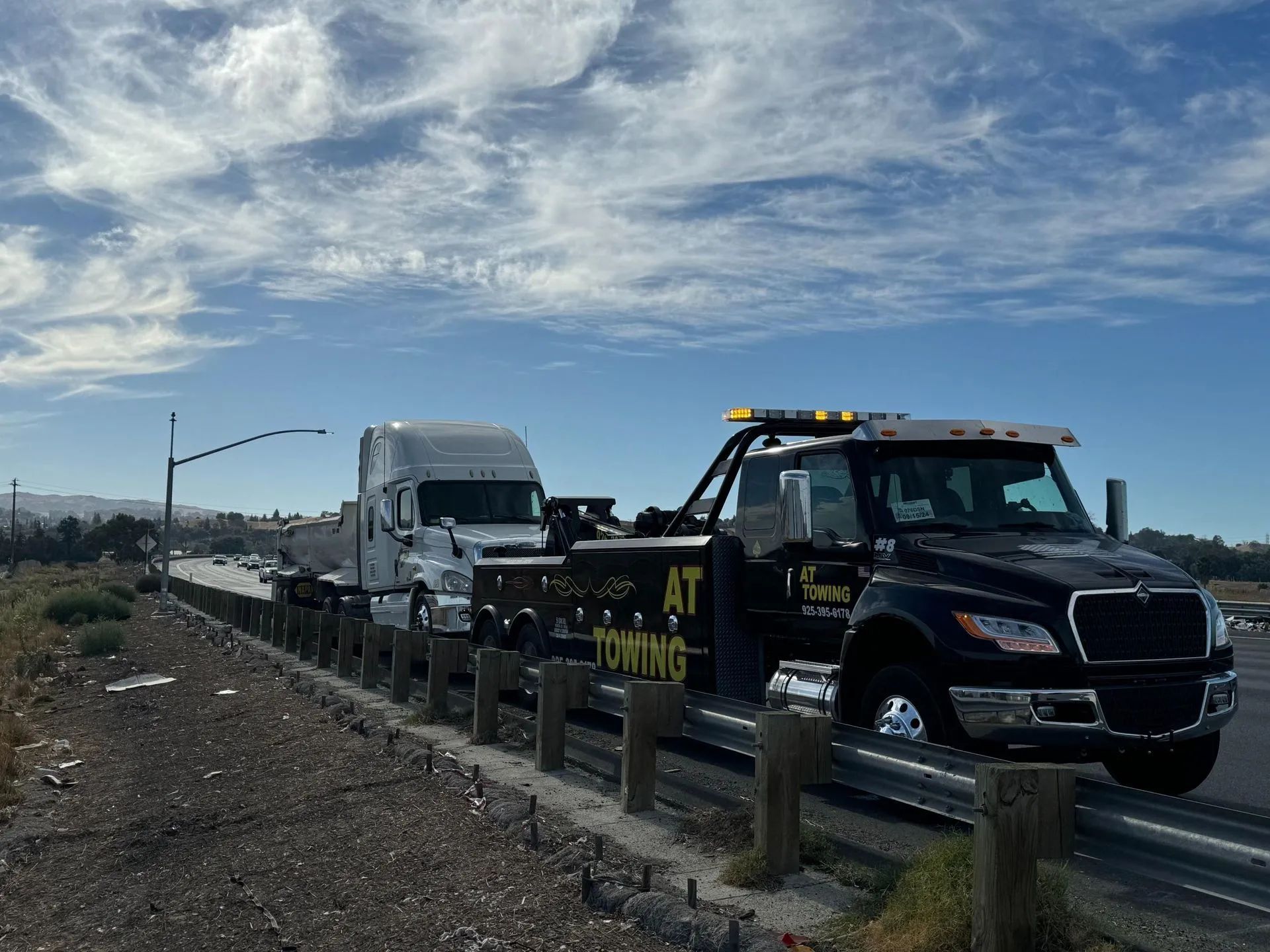 A black tow truck with the words AT TOWING pulls a white semi-truck along a highway shoulder under a blue, cloudy sky.