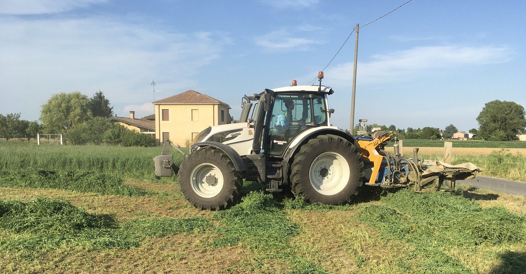 Un trattore sta tagliando l'erba in un campo con una casa sullo sfondo.