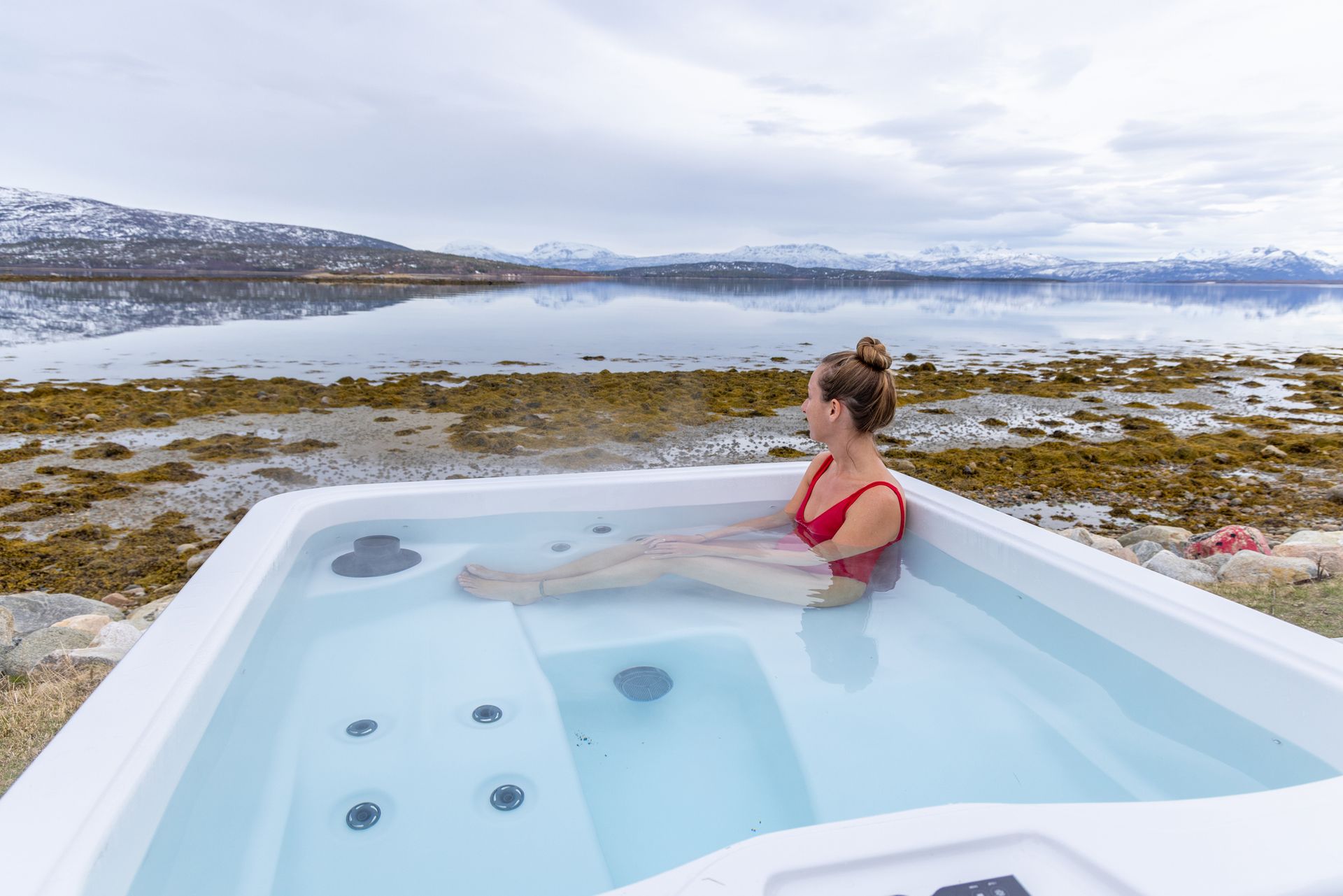 Woman in red swimsuit soaks in hot tub, admiring snowy mountains by a peaceful lake. Woman in red swimsuit soaks in hot tub, admiring snowy mountains by a peaceful lake.
