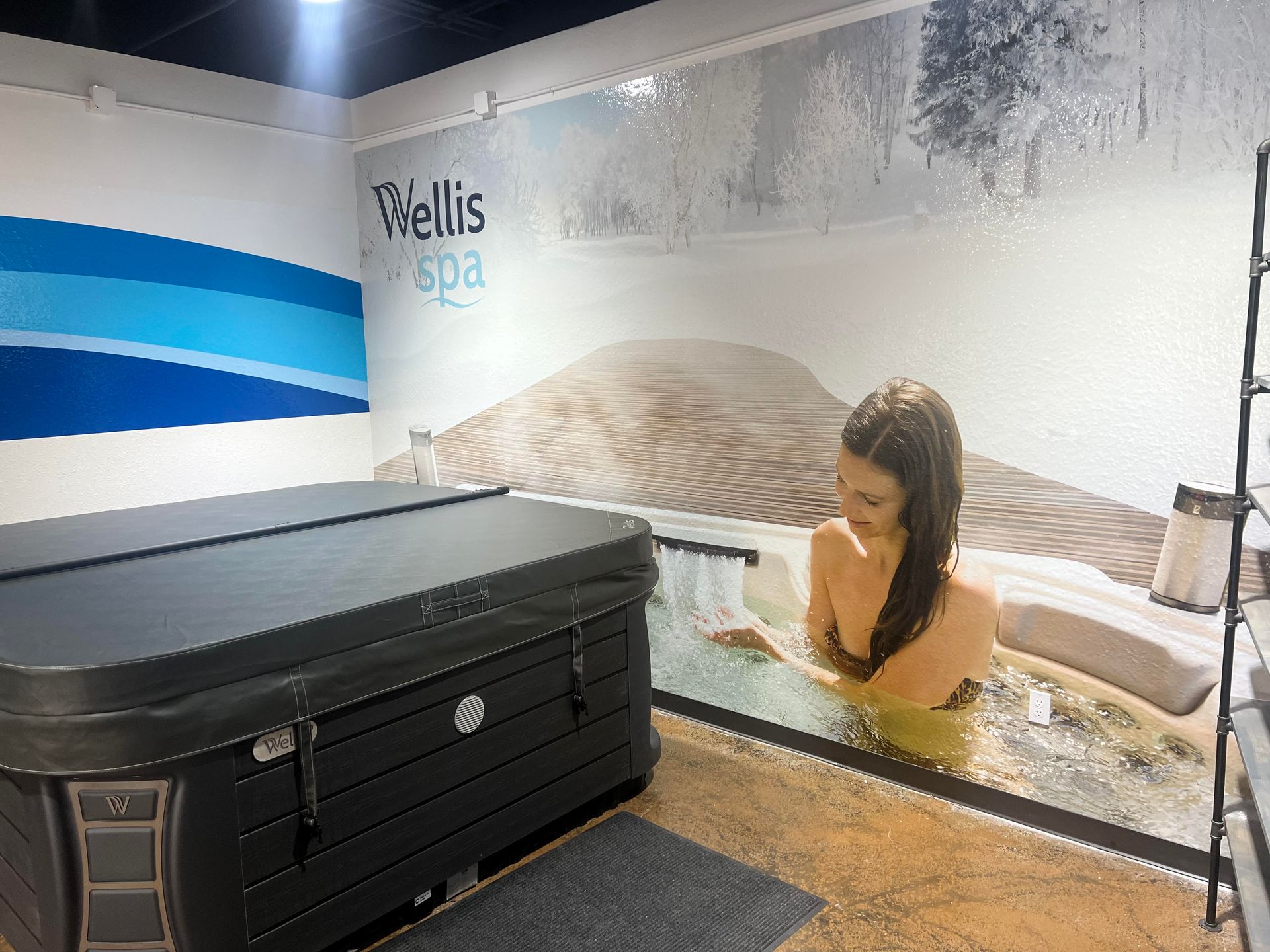 A woman in a hot tub display with a closed hot tub cover, Wellis Spa logo, and a wall mural.