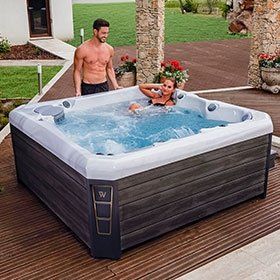 A man and woman in a hot tub on a wooden deck; stone columns in the background.
