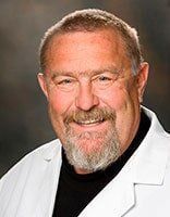 A man with a beard is wearing a white lab coat and smiling.