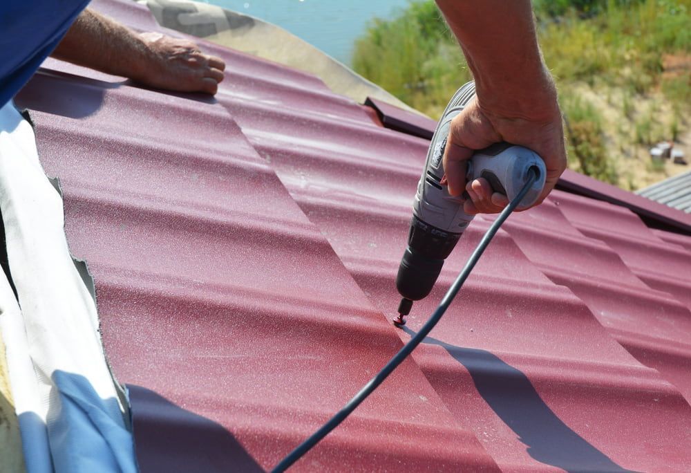 A Man is Using a Drill on a Red Roof — Woodger's Metal Roofing Specialists Pty Ltd in South West Rocks, NSW