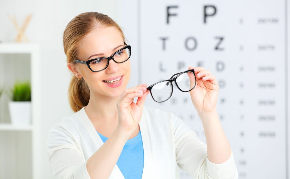 A Woman Is Holding a Pair of Glasses in Front of An Eye Chart — John Williams Optometrist in Grafton, NSW