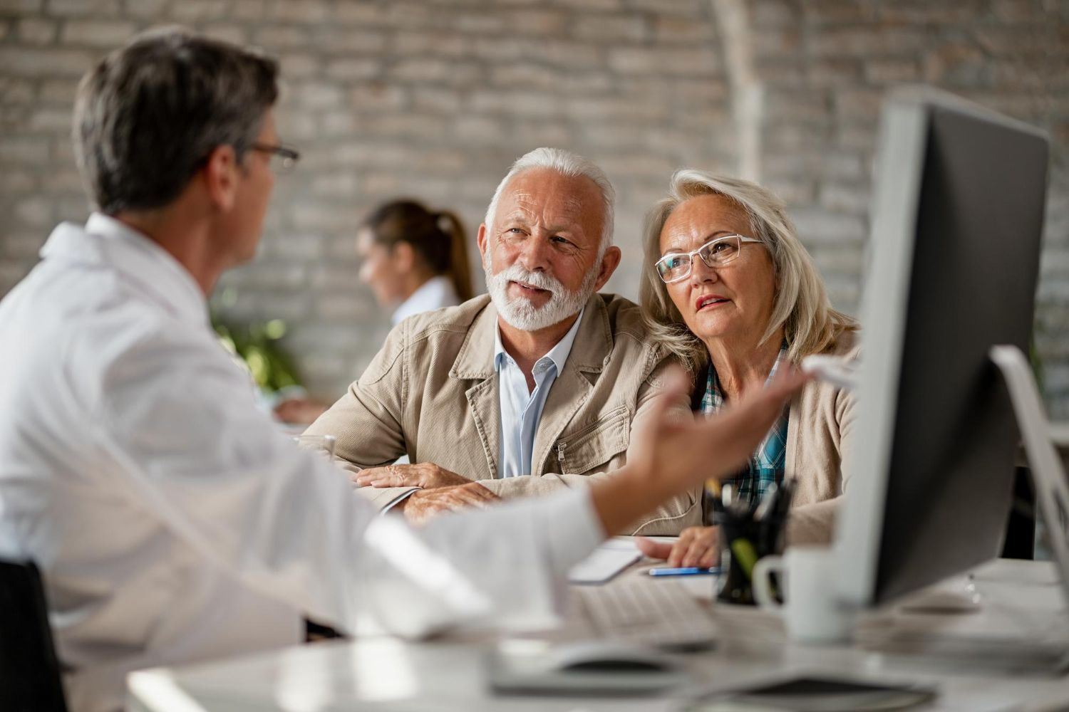 Doctor speaking with two patients in a clinic, seated at a table with a computer monitor in the foreground.