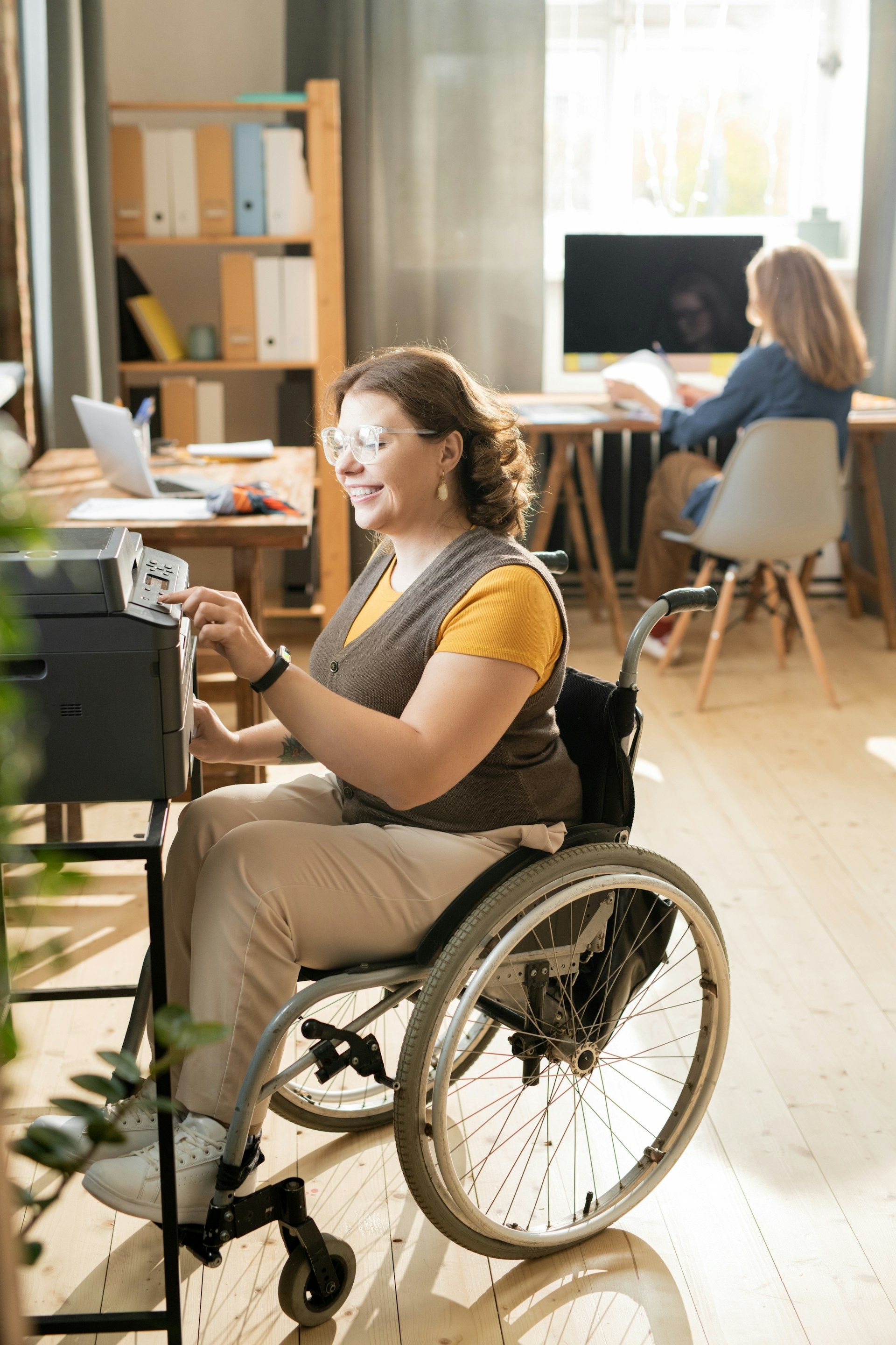 Woman in a wheelchair working at a computer in a bright office with another person in the background