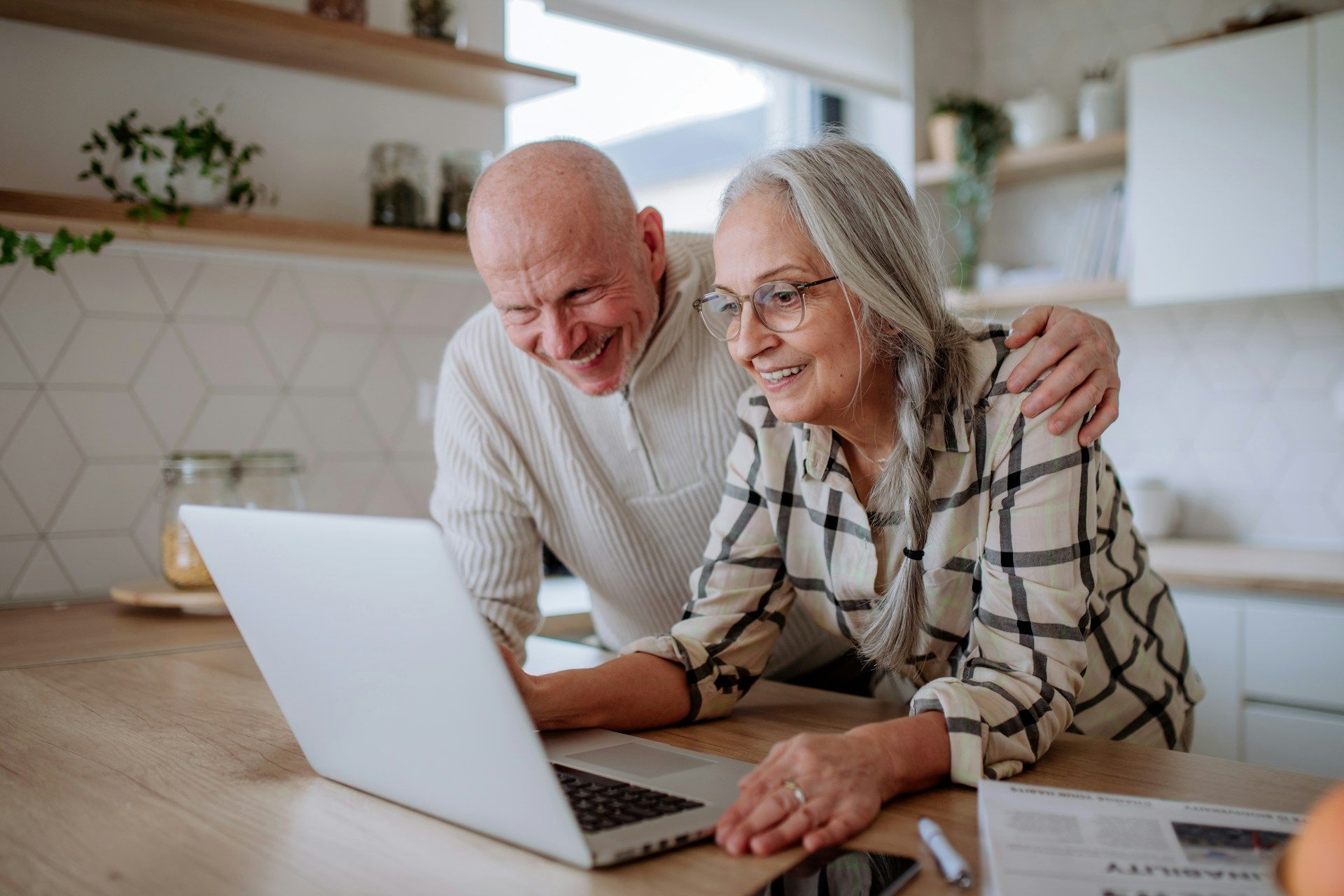 Two smiling people using a laptop at a kitchen table, with one pointing at the screen