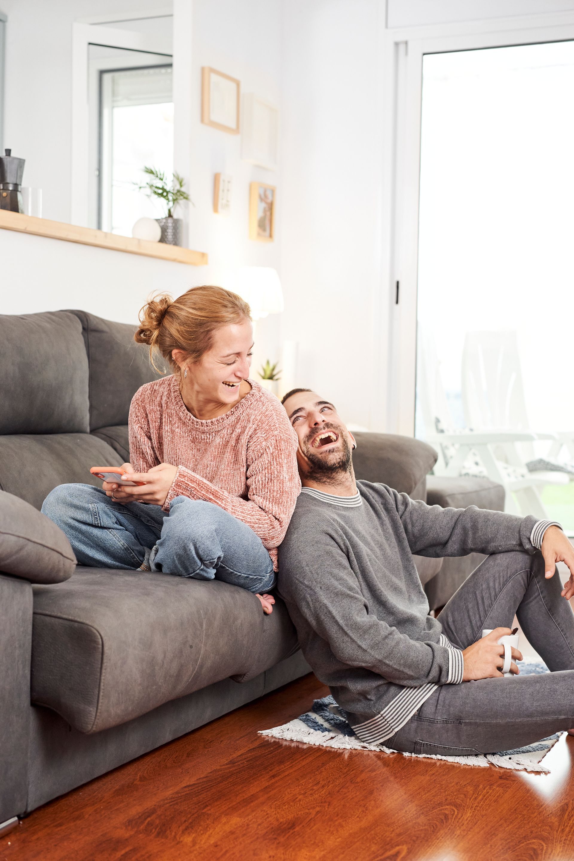 Couple laughing on a gray couch in a bright living room, one holding a tablet.