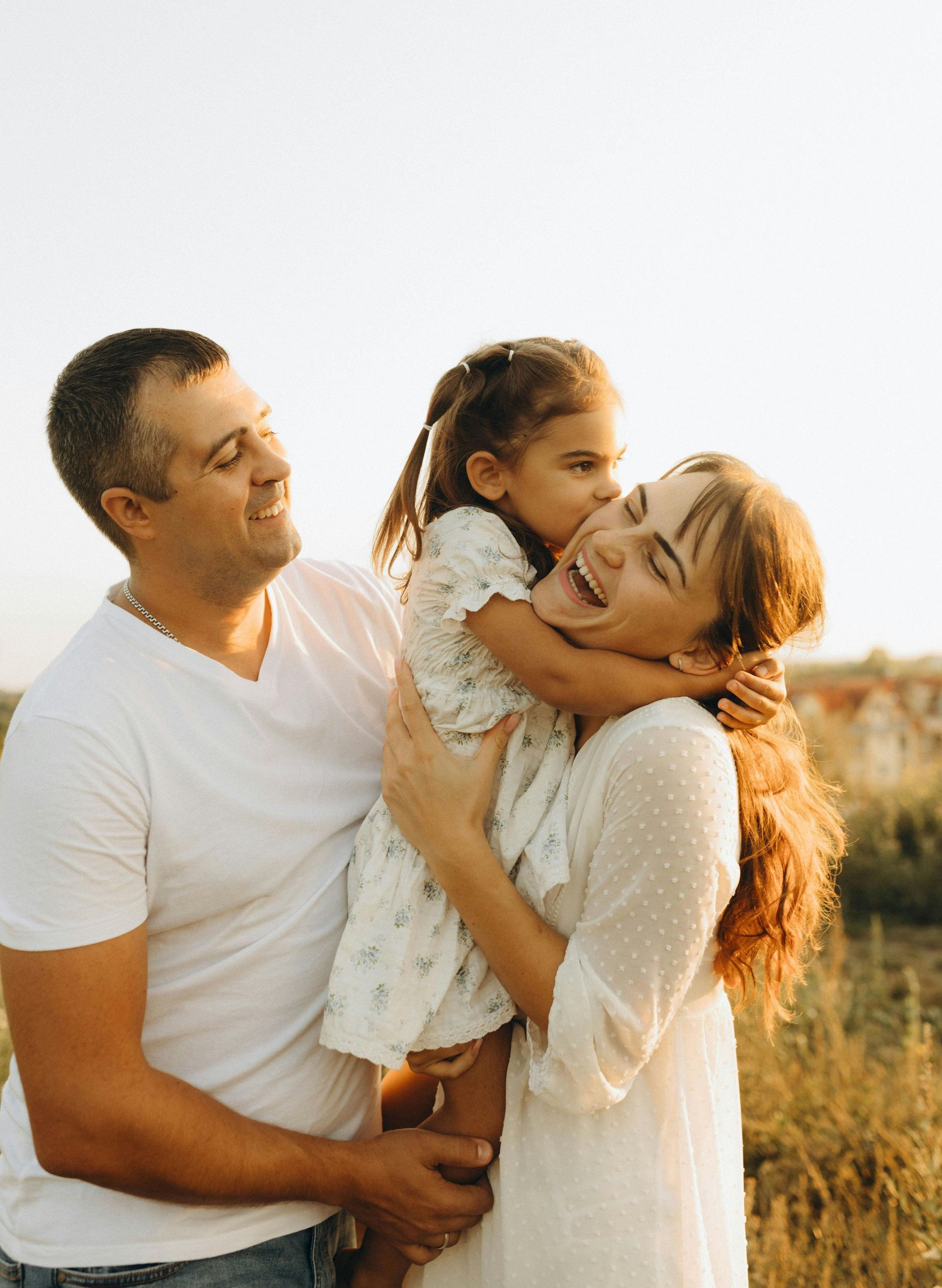 Family hugging outdoors at sunset, with a child held between two smiling adults.
