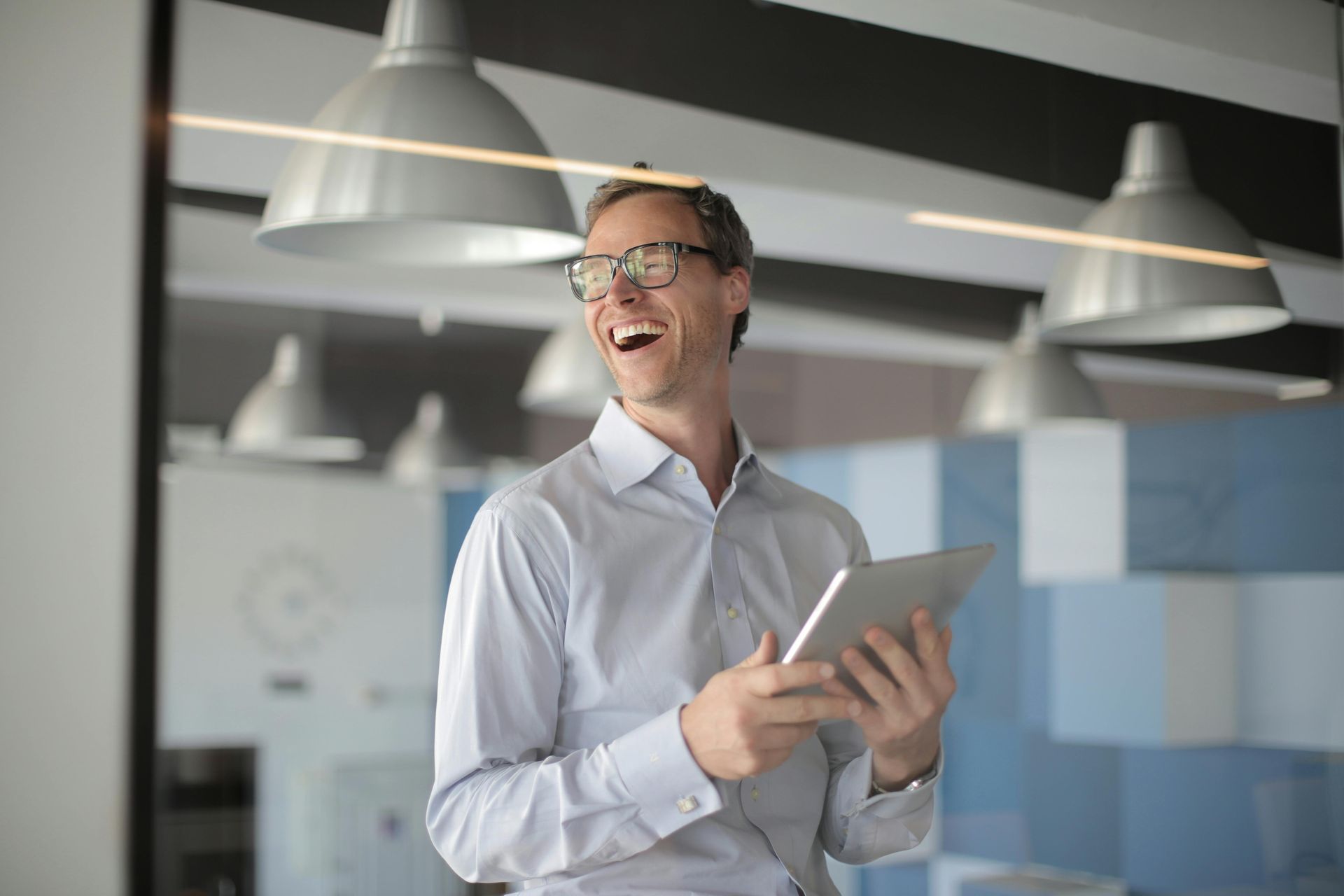 Smiling person in glasses holding a tablet in a modern office with hanging pendant lights.