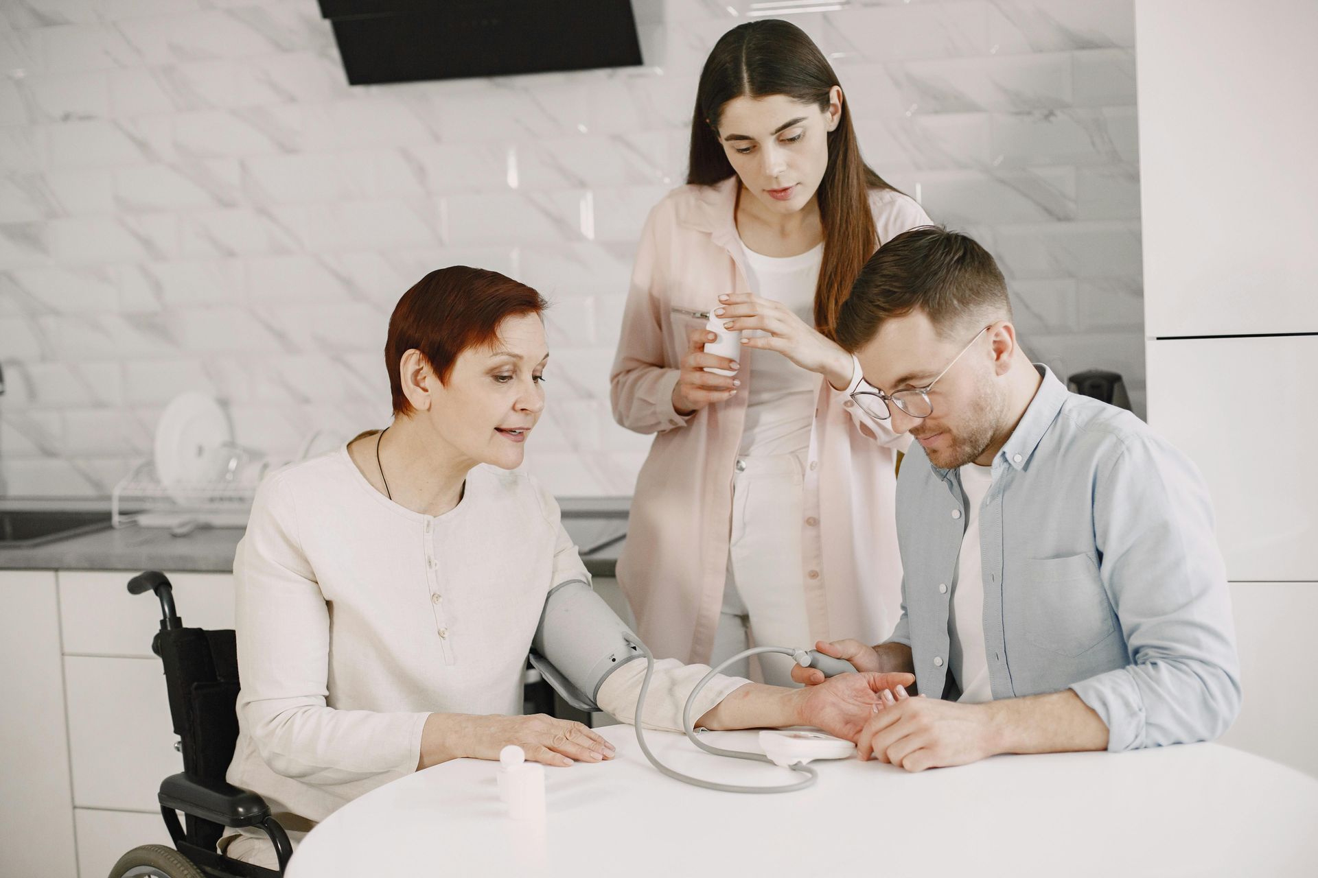 Three people in a bright kitchen examine a patient’s hand during a medical checkup