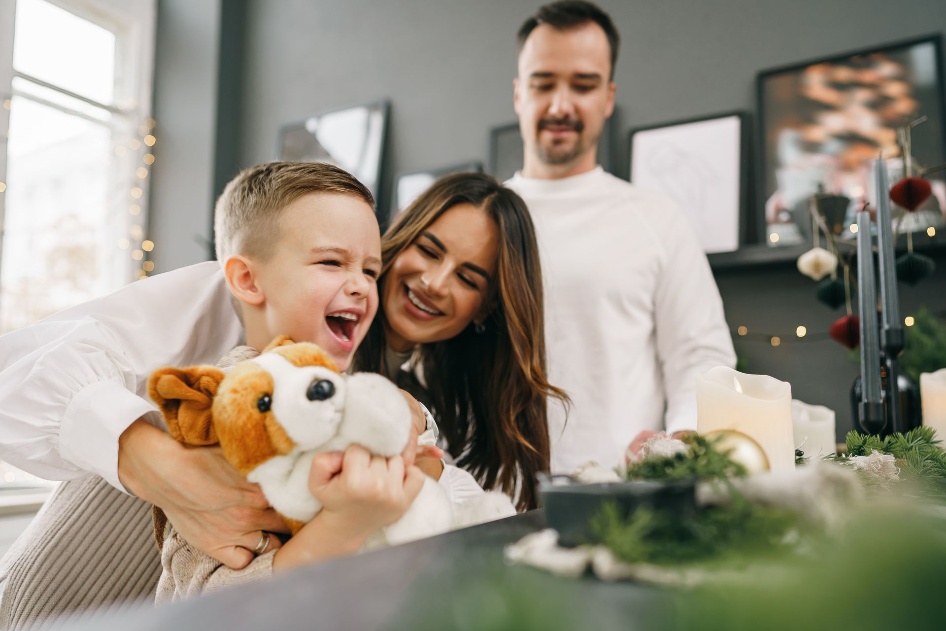 Woman and child holding stuffed animals, with a man standing behind them in a bright living room.