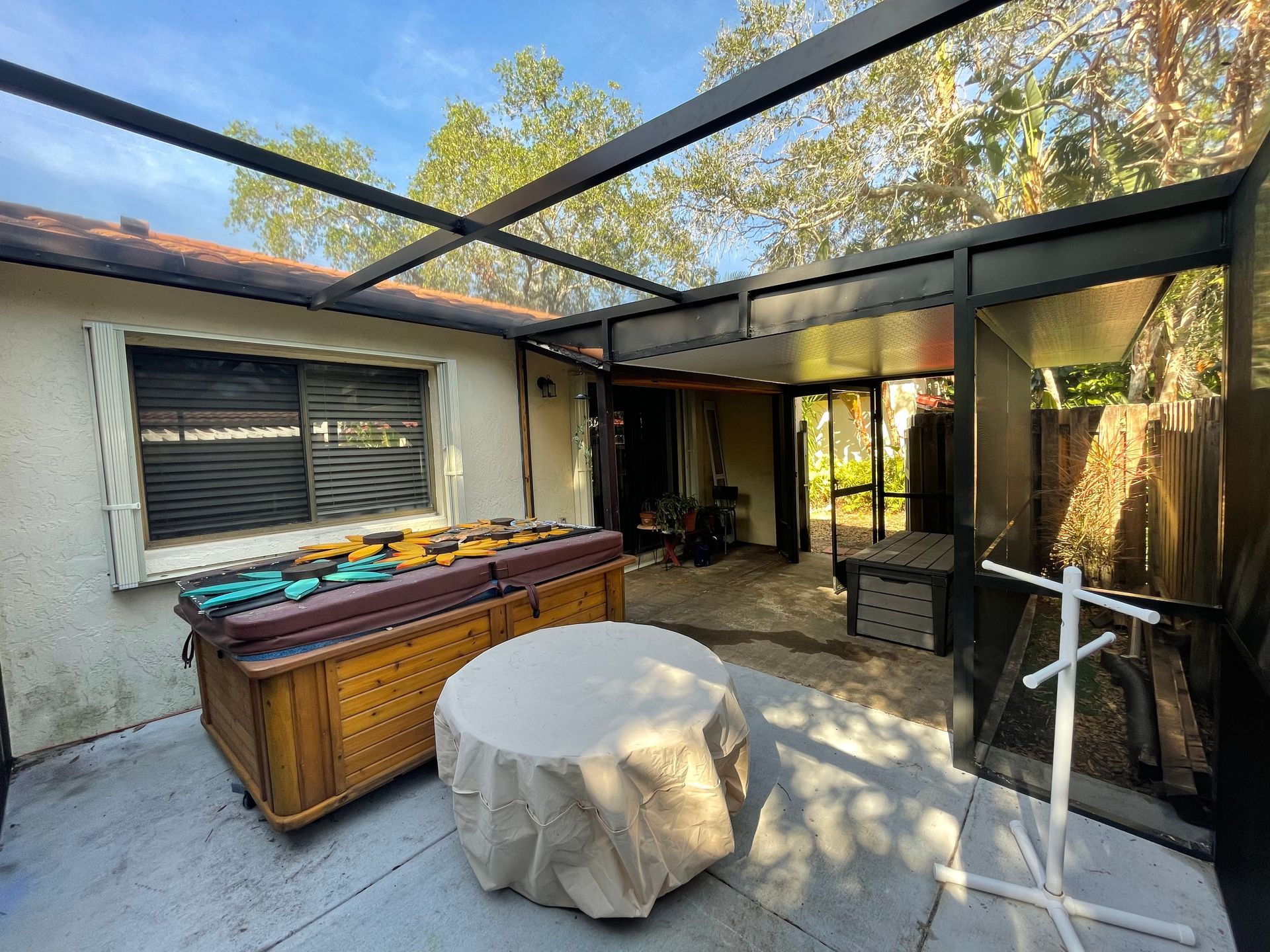 A backyard patio enclosed by a screened-in frame features a hot tub, a covered round table, and a view of trees.