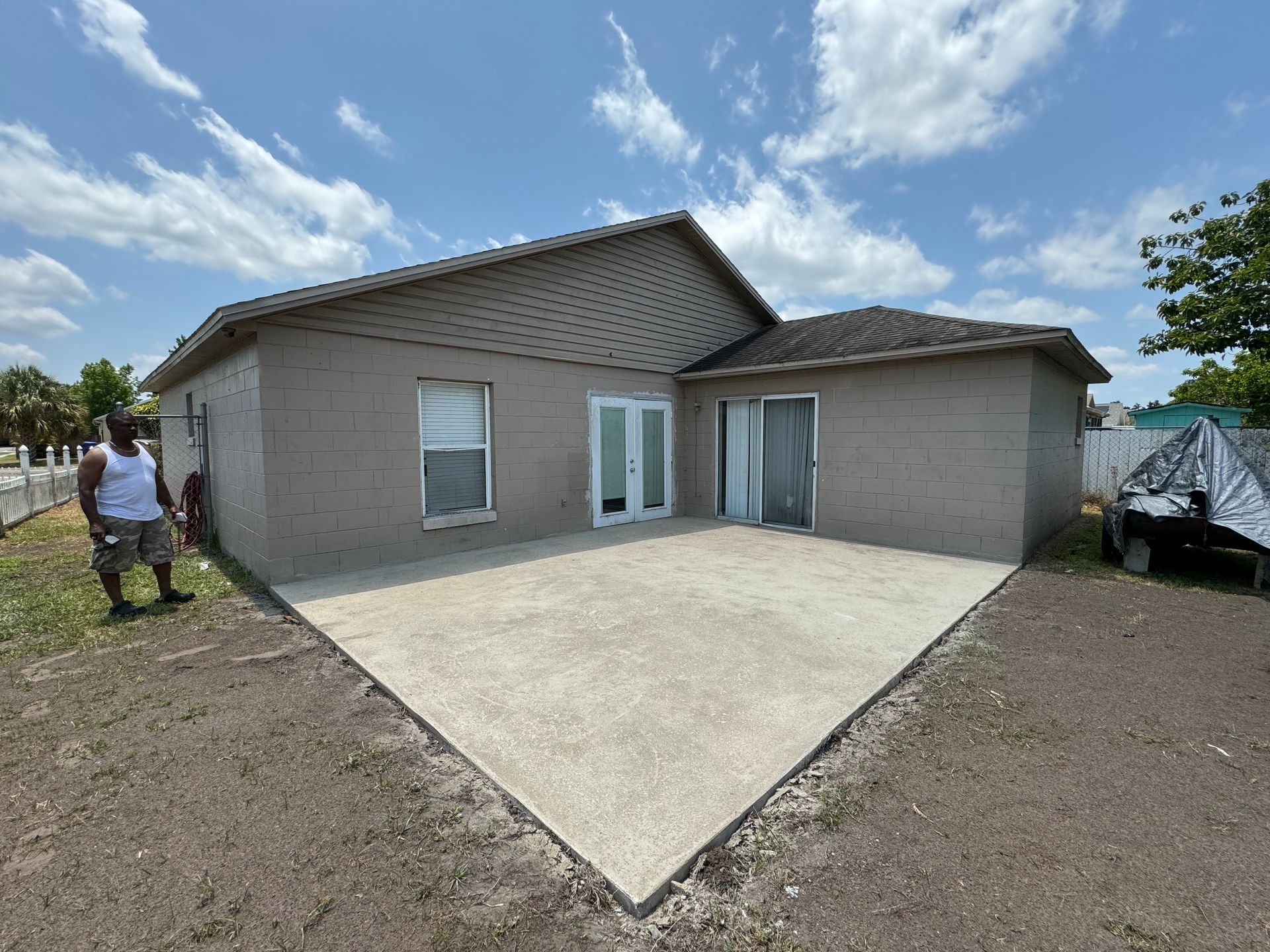 A person stands on the grass beside a newly poured concrete patio attached to the rear of a beige, single-story house.