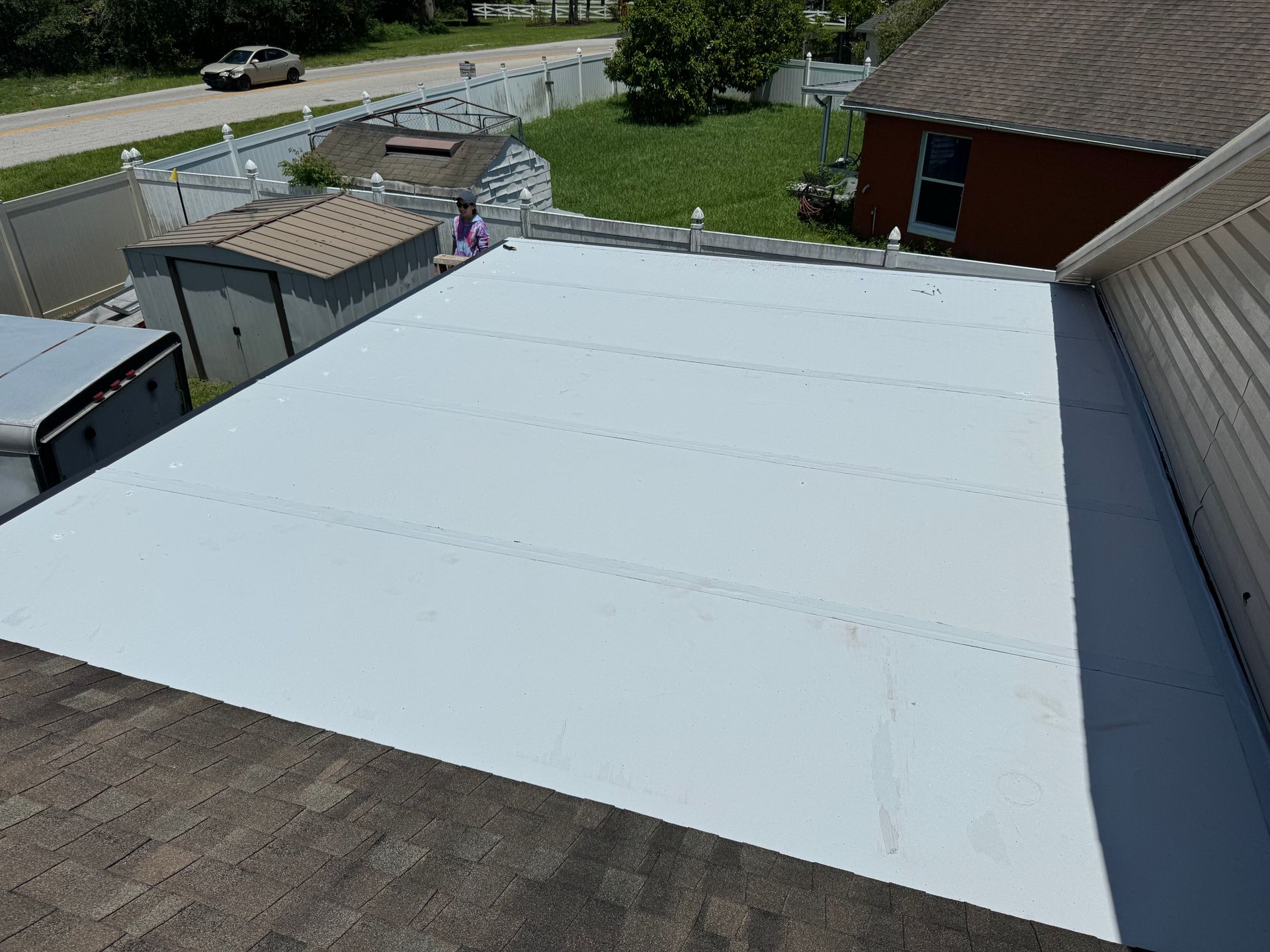 A white flat roof surface viewed from above, with sections of shingled roofing and a house exterior visible nearby.