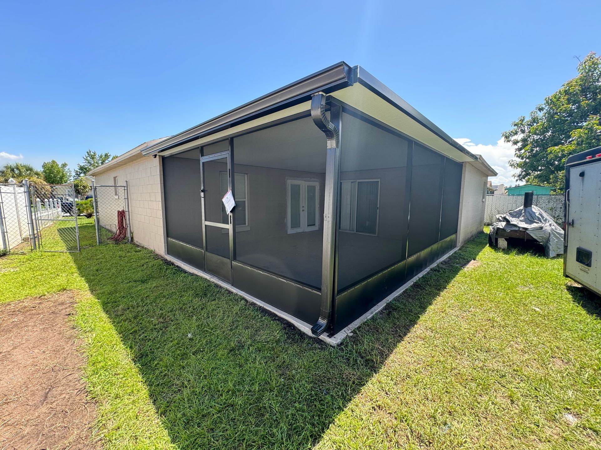 A corner view of a beige house with a black-framed screen enclosure attached to the back, set on a bright, sunny lawn.