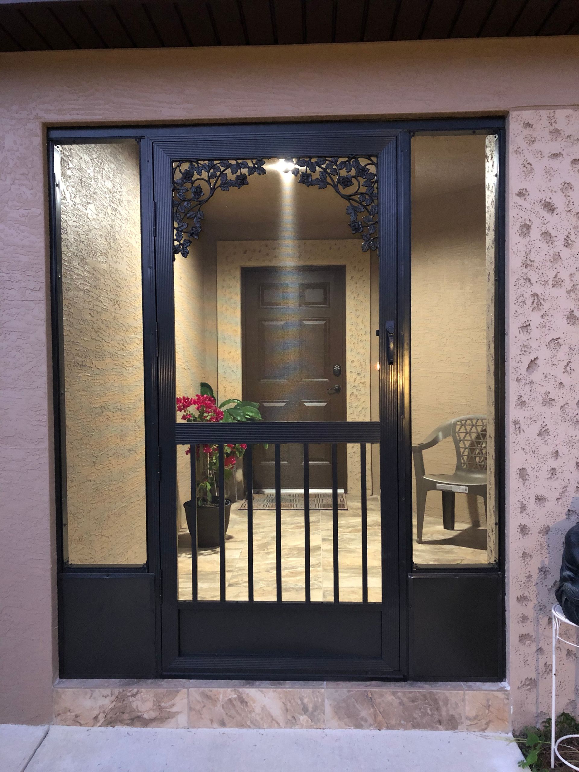 Black security storm door with decorative scrollwork, flanked by vertical glass panels on a light-colored building facade.