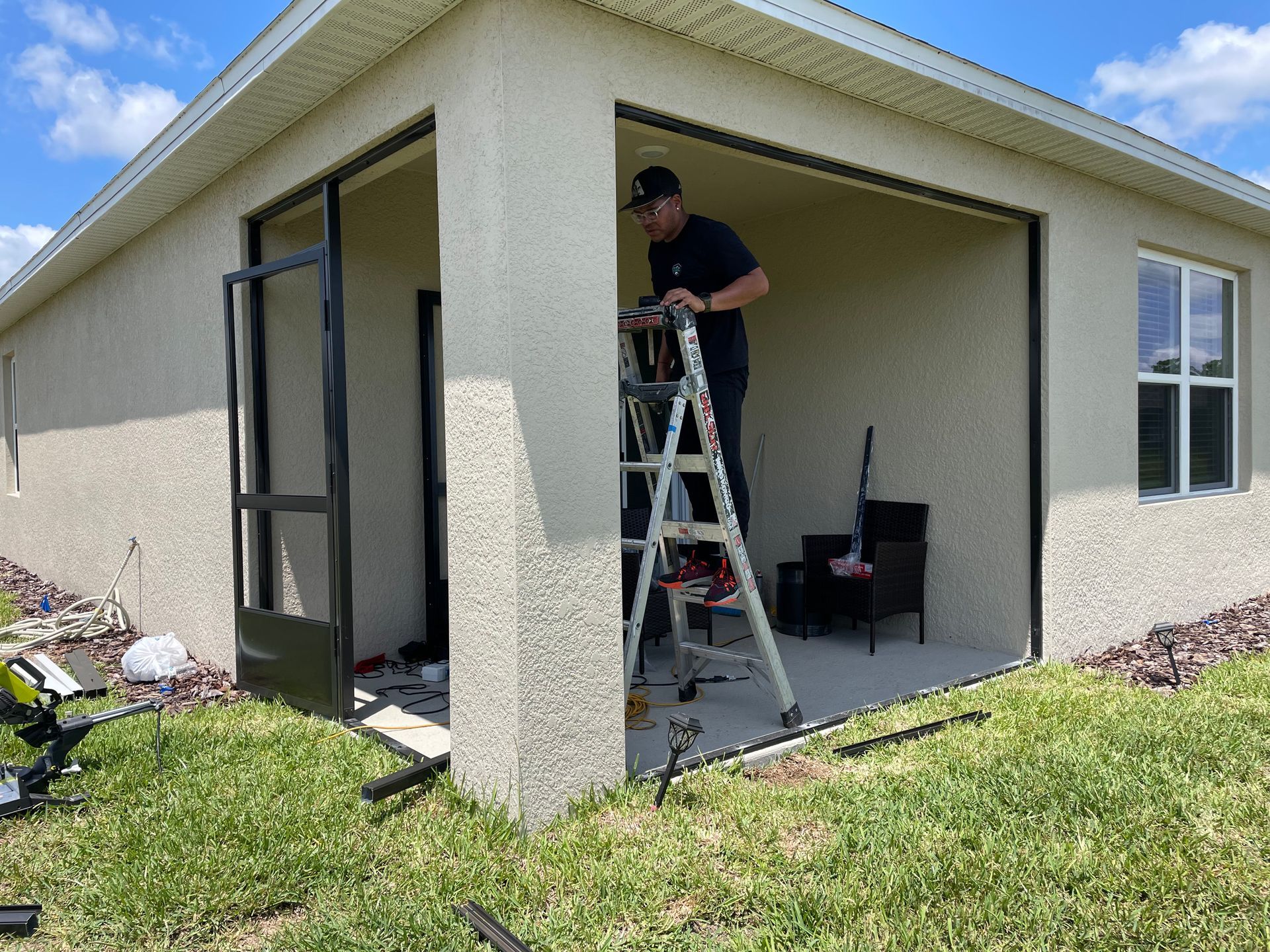 A worker on a ladder installs a screen door on a patio enclosure outside a light-colored stucco house.