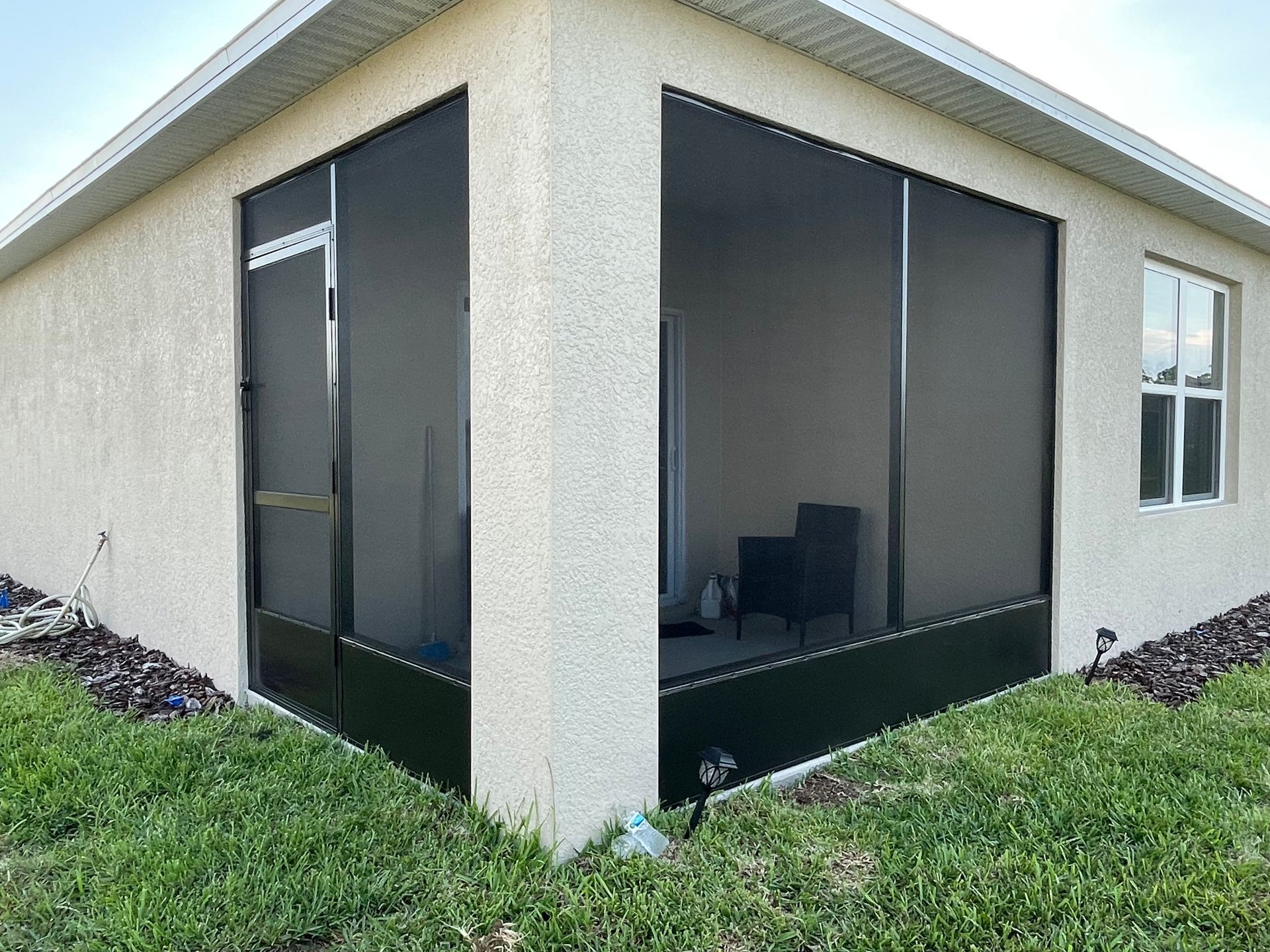 Corner view of a beige stucco house featuring an enclosed screened-in patio with black framing and kick plates.