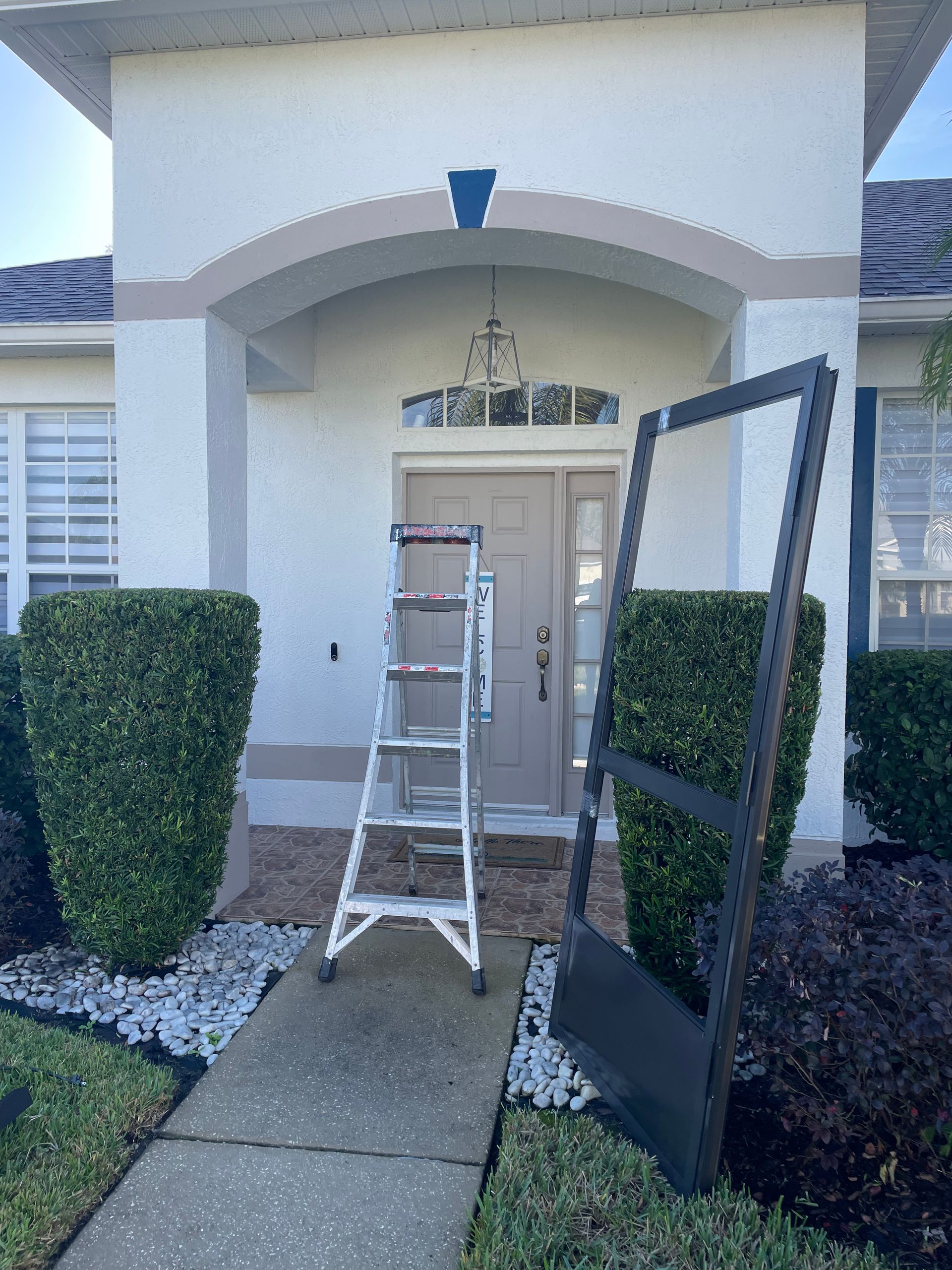 A metal ladder stands on a concrete path before a front door, next to a detached dark gray storm door frame.