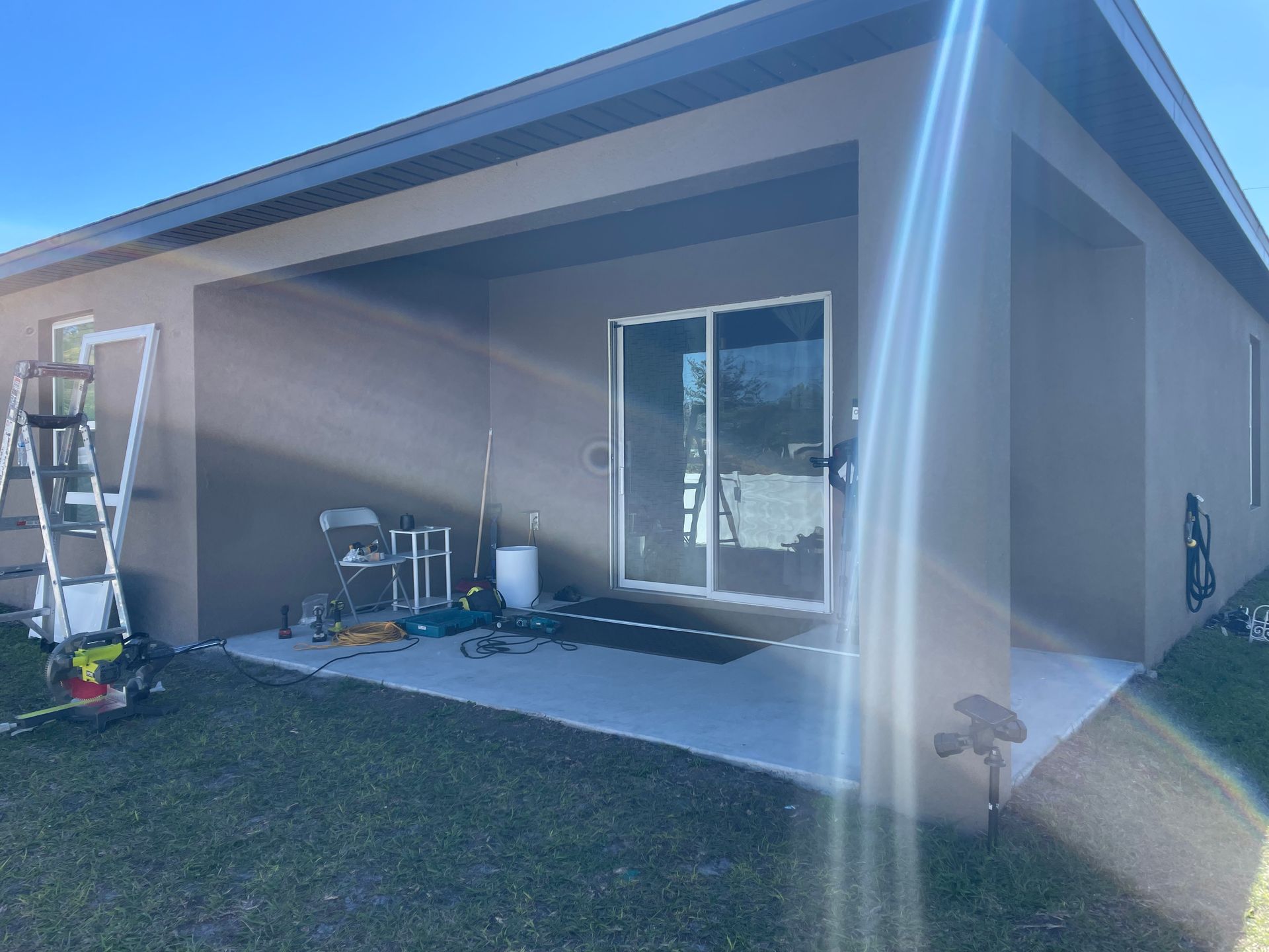 A tan house exterior with a covered concrete patio, sliding glass doors, and construction tools on the ground.