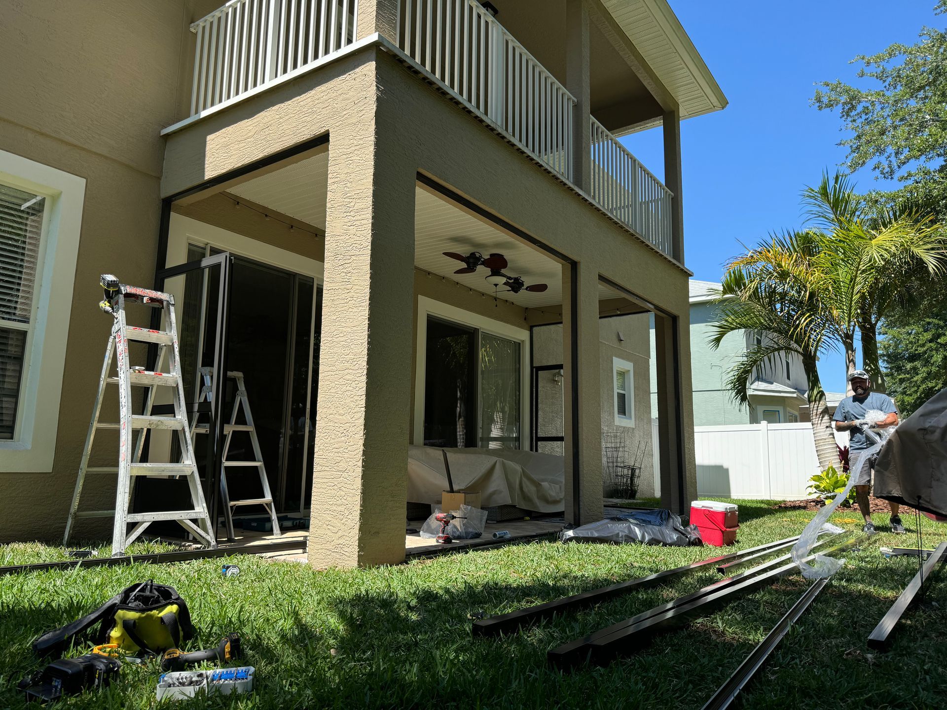 A person stands near a patio under construction, with a ladder, tools, and framing materials on the grassy lawn.