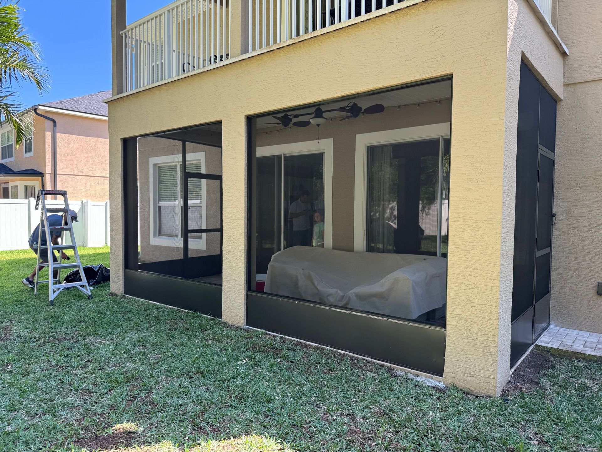 A worker sets up a ladder outside a tan house with a screened-in ground-floor porch featuring a covered piece of furniture.