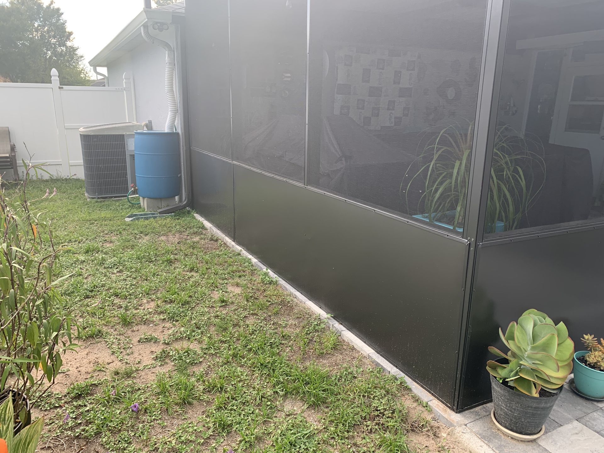 A black screen enclosure exterior in a backyard, featuring a blue rain barrel and potted plants on a patio.