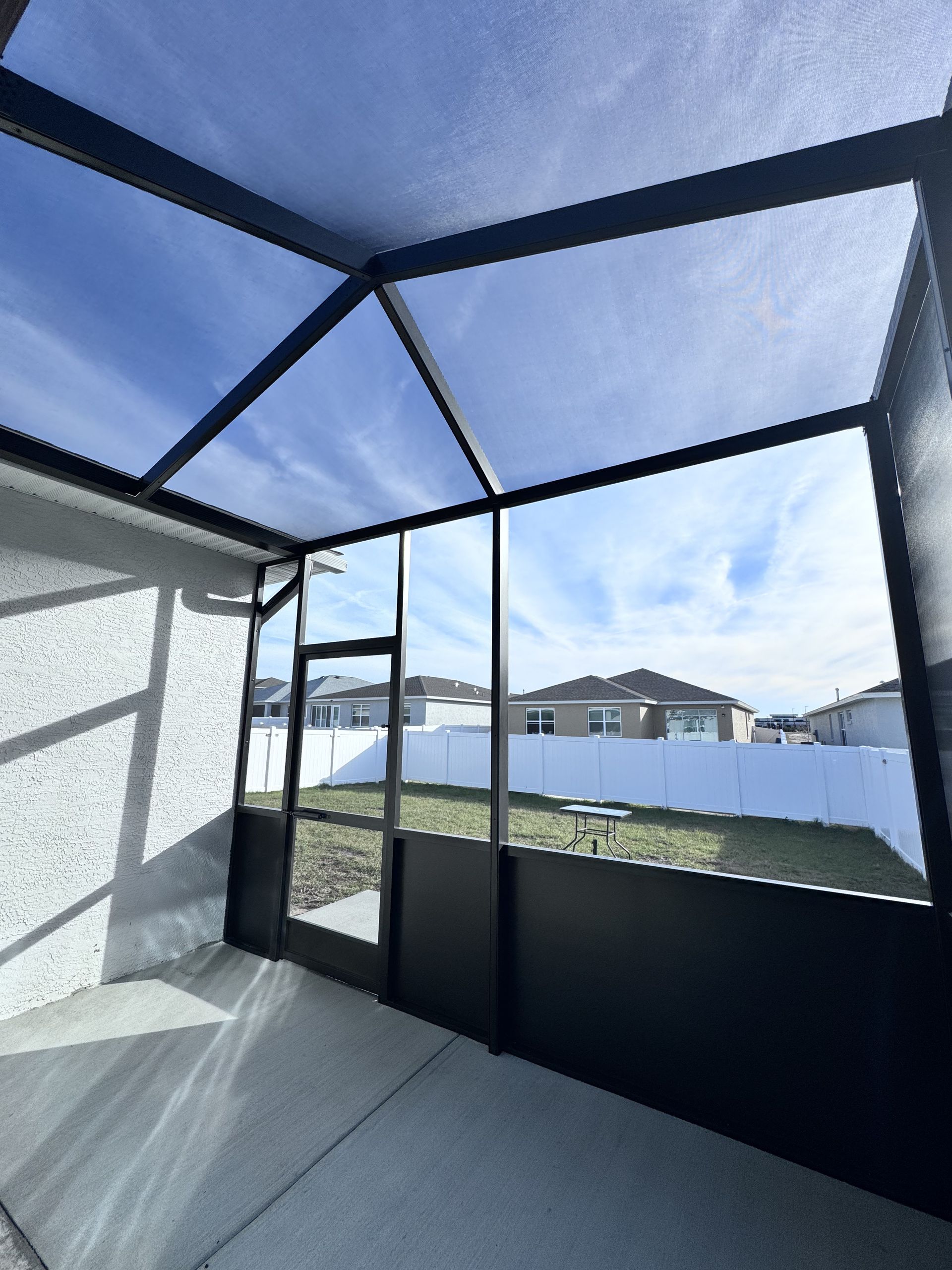 A screened-in patio overlooking a fenced backyard with neighboring houses under a blue sky.