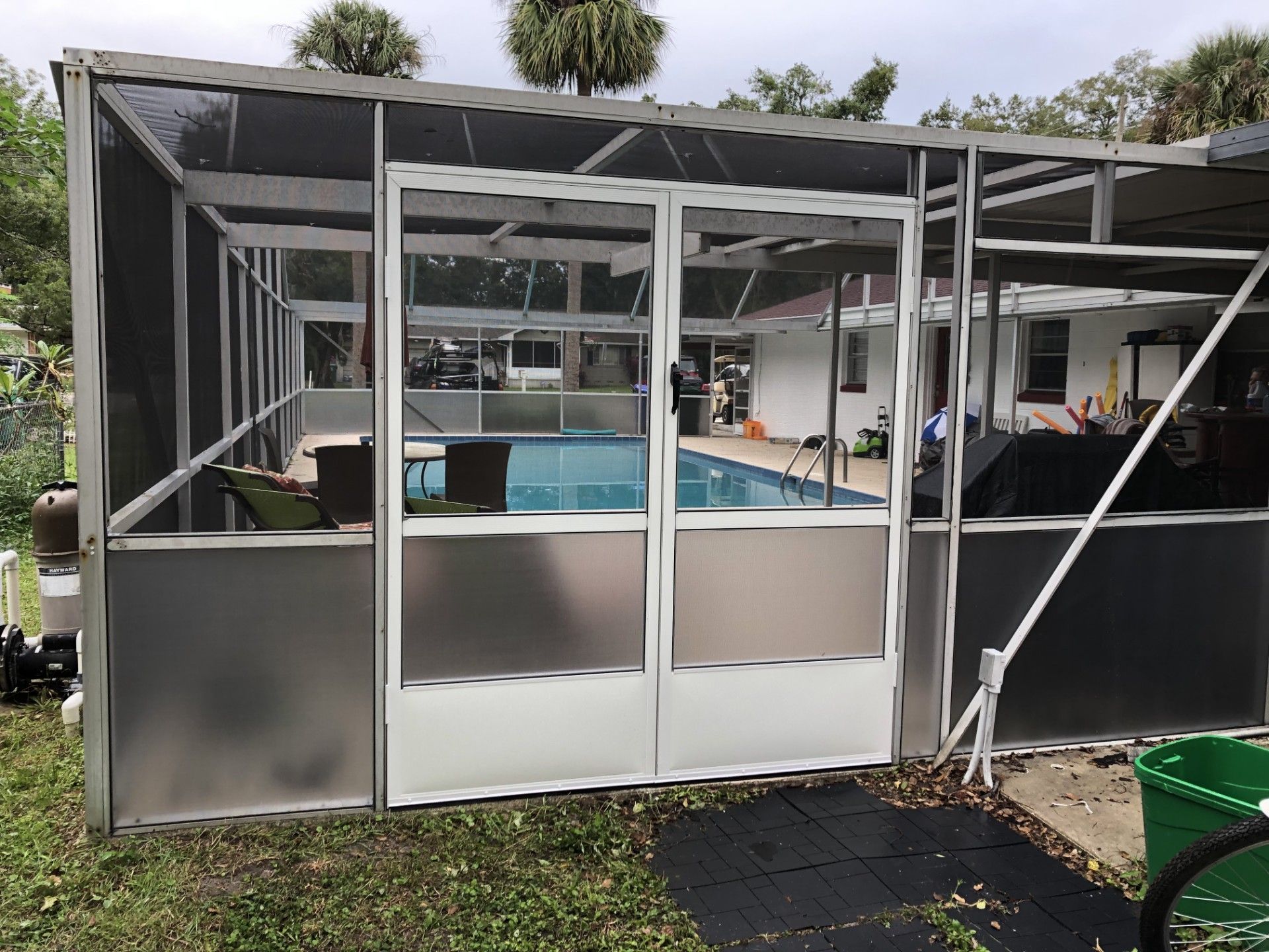 White-framed swimming pool screen enclosure with a double door and metal kickplates in a backyard setting.
