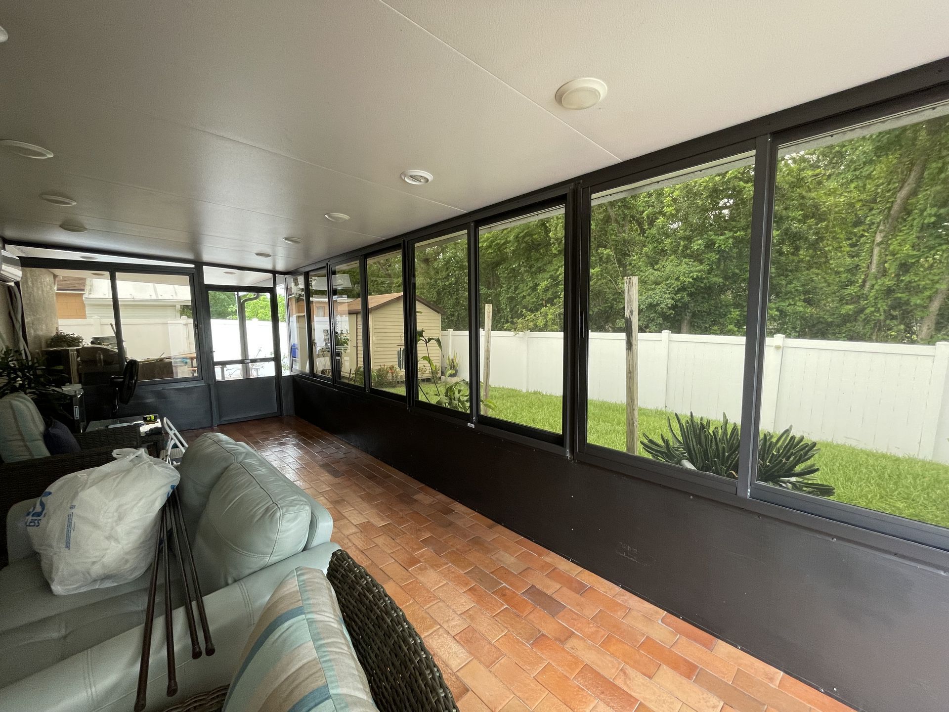 A sunroom with a brick floor, light-colored furniture, and a large bank of windows looking out at a grassy backyard.