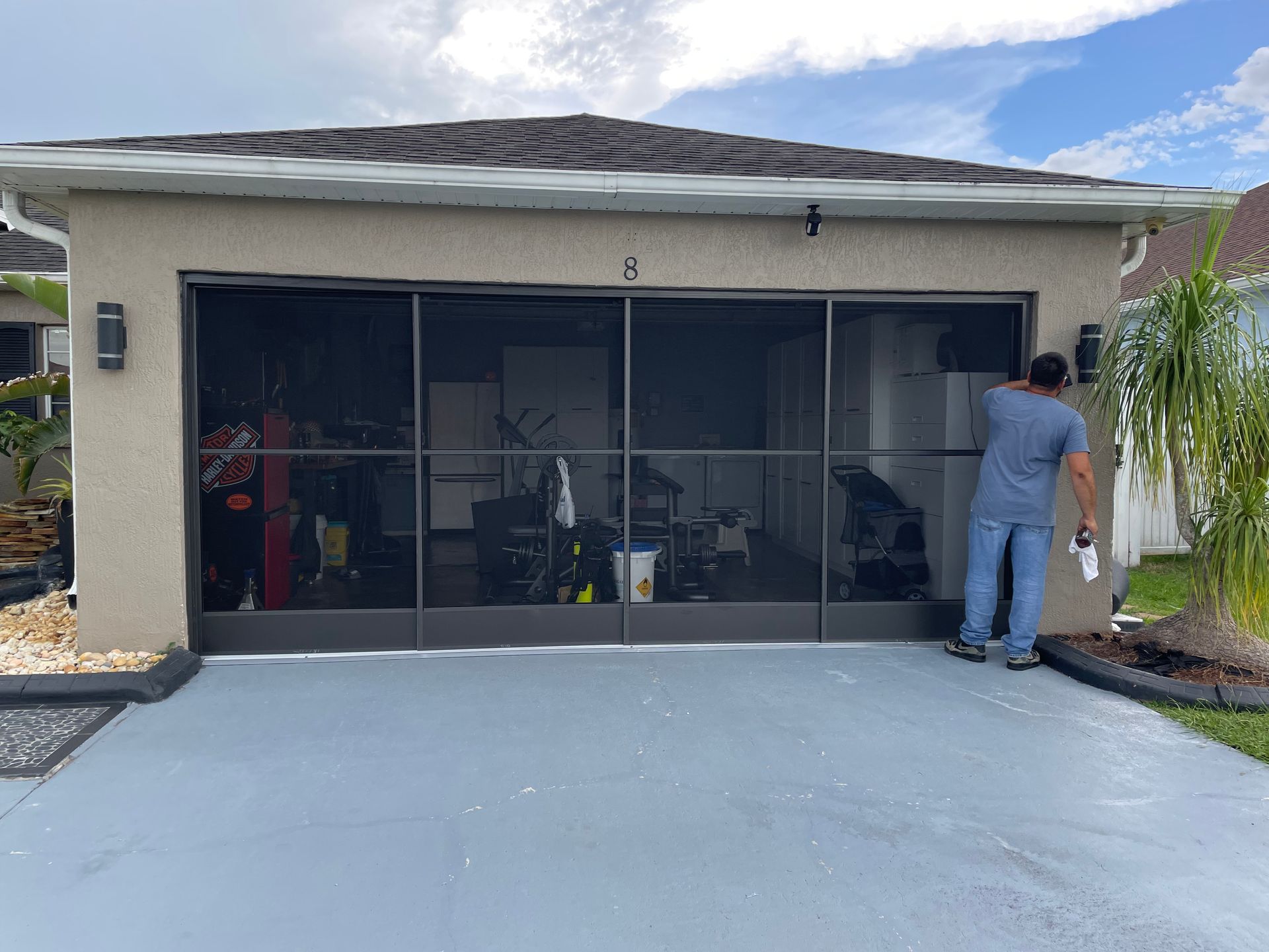 A person installing a dark, multi-panel garage screen door on a tan stucco house with a gray painted driveway.