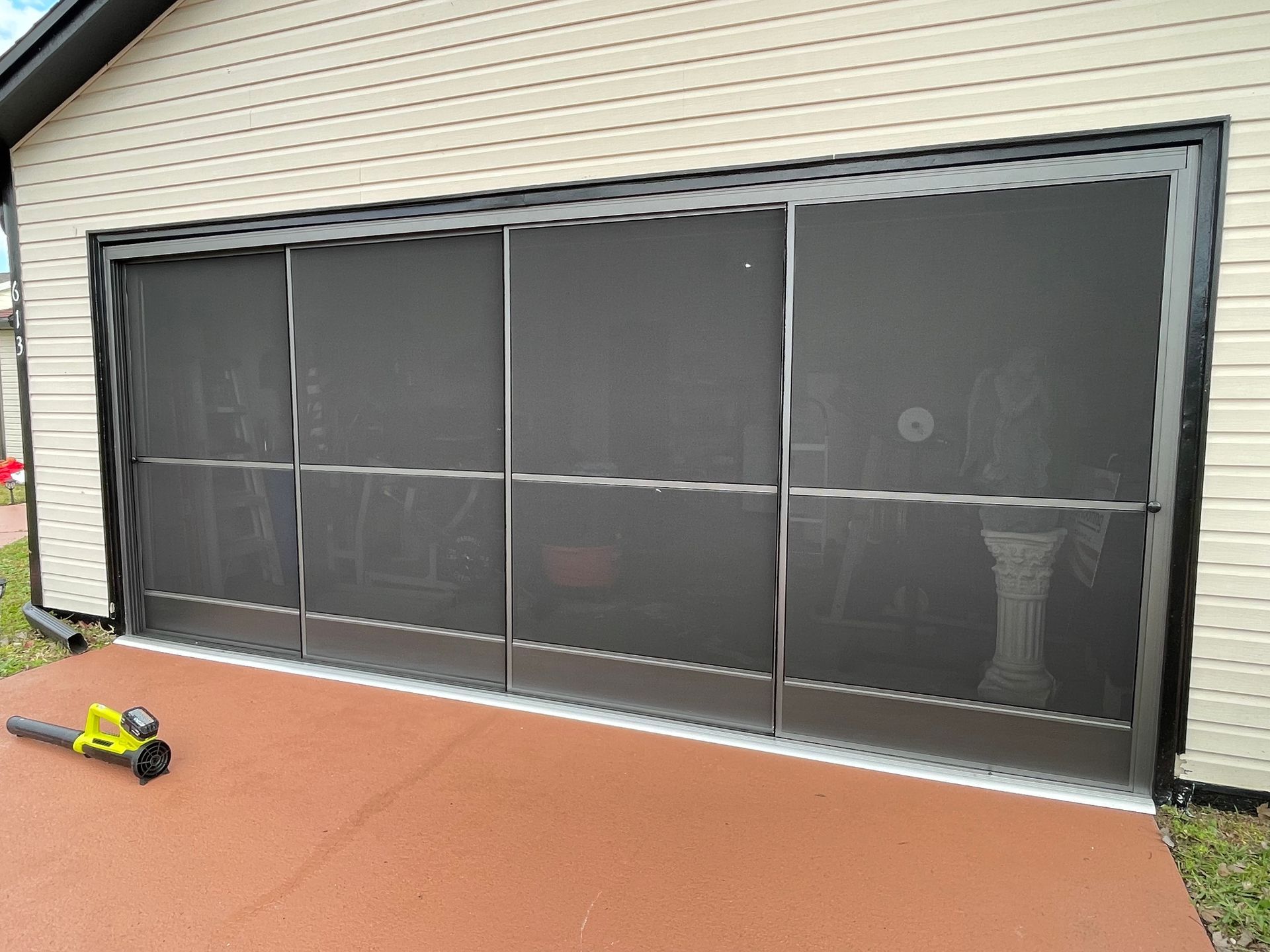 A dark, four-panel screen door installed on a garage opening with tan siding and a small leaf blower on the patio.