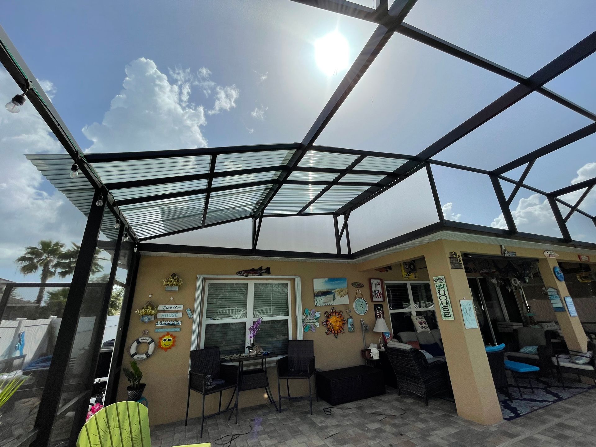 A sunny patio area enclosed by a screened-in roof, featuring outdoor furniture, wall decorations, and a palm tree nearby.