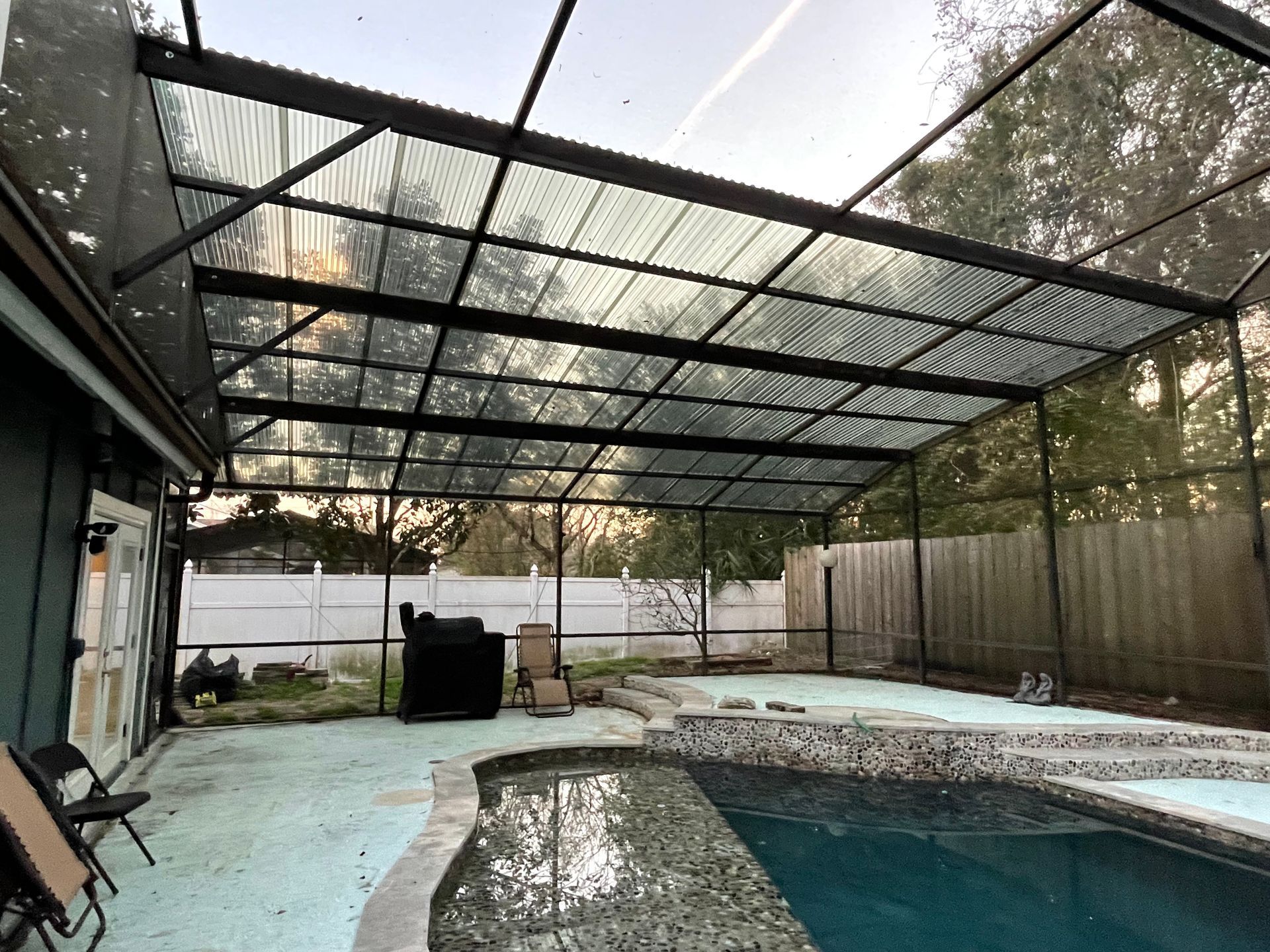 A backyard swimming pool area with a covered screen enclosure, light-colored patio deck, and a white privacy fence.