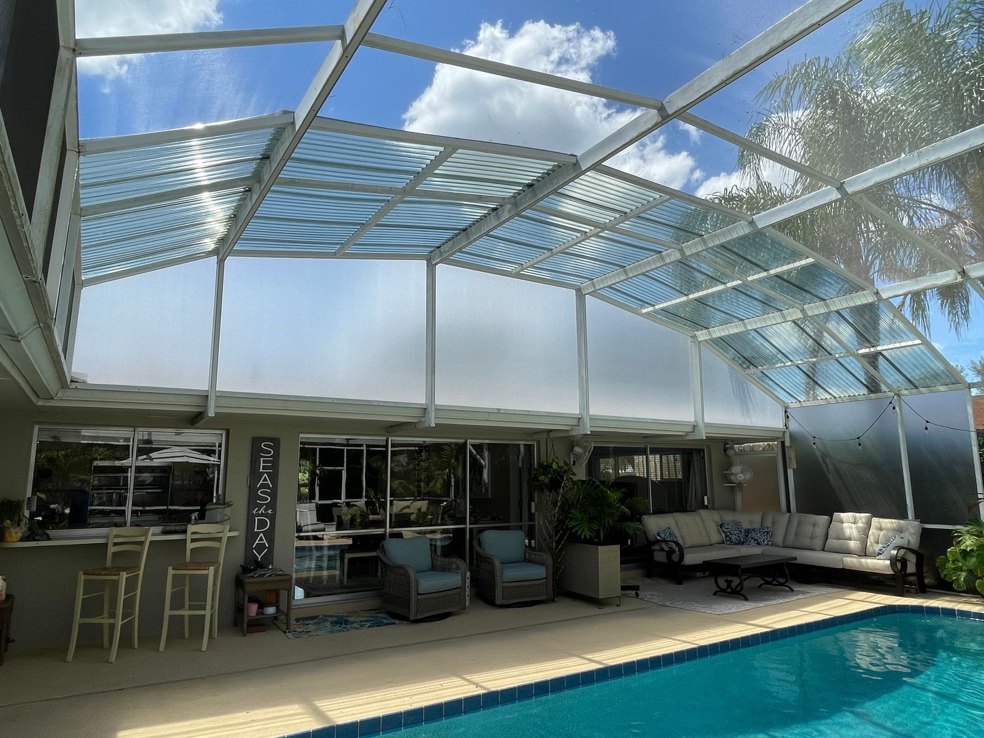 A covered patio featuring a swimming pool, outdoor seating, and a white metal screened-in roof under a sunny blue sky.