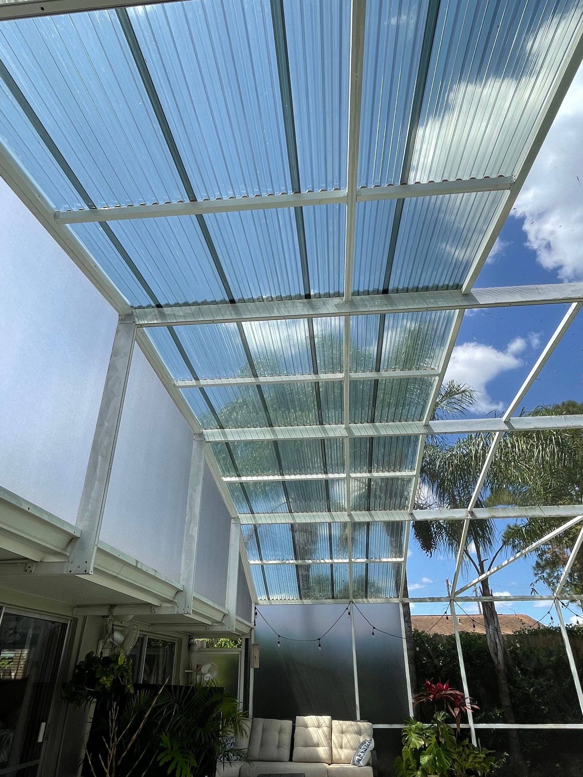 A view from under a white-framed patio screen enclosure with a clear corrugated roof against a blue sky with clouds.