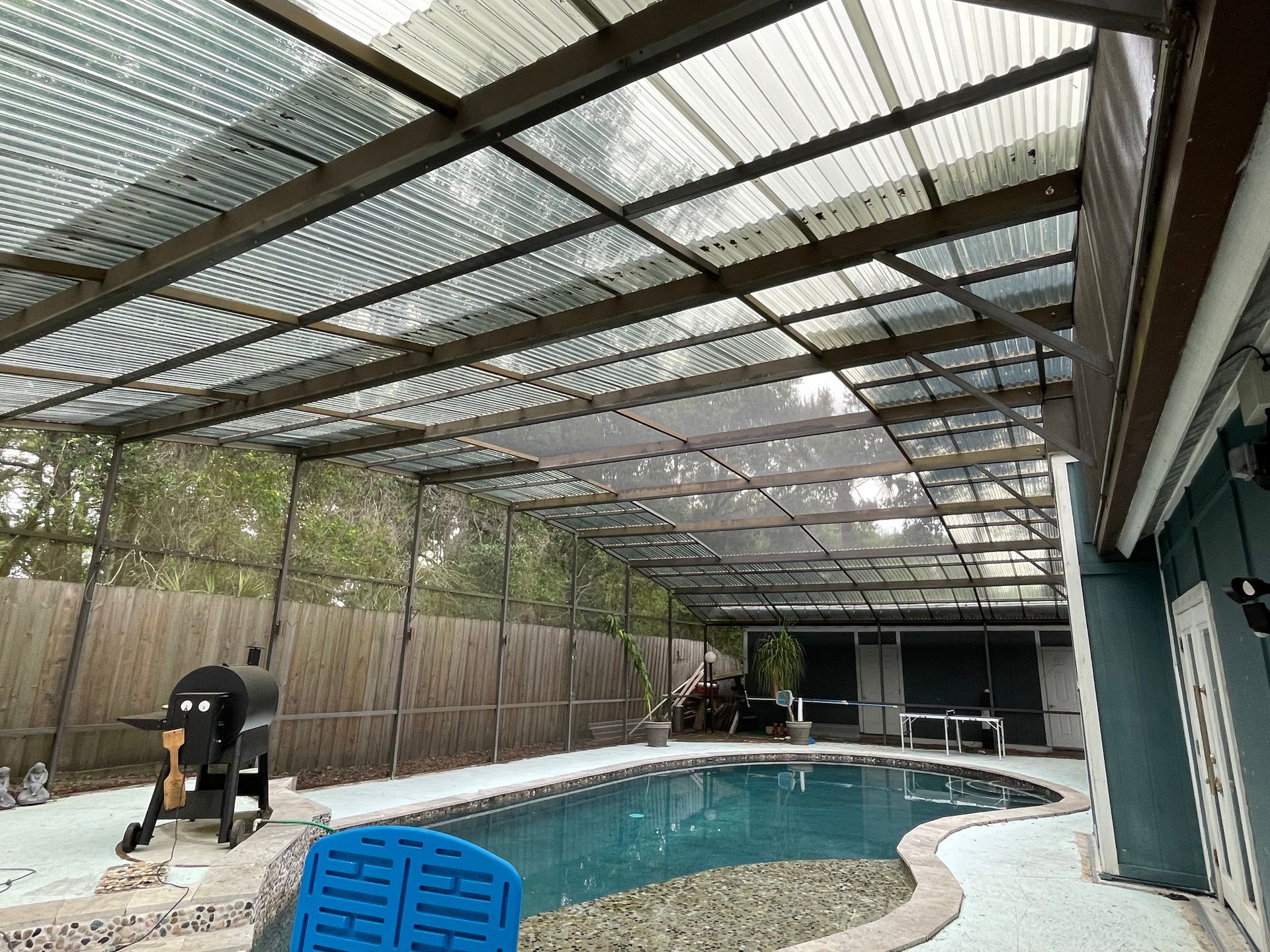 A backyard pool enclosed by a wooden structure with a clear corrugated roof, featuring a grill and patio area.