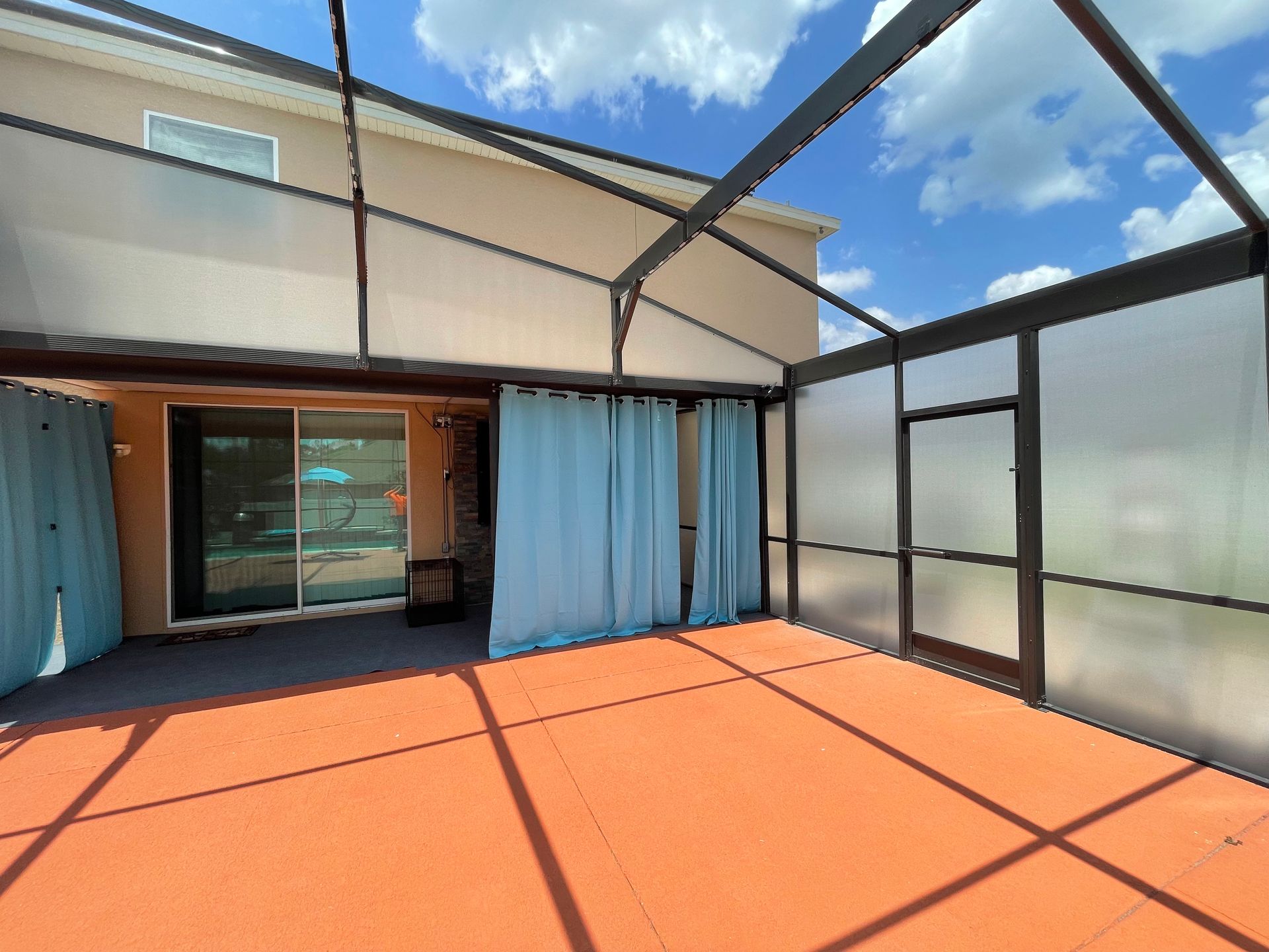 A covered patio with light-blue curtains, orange flooring, and a sliding glass door leading into a house.