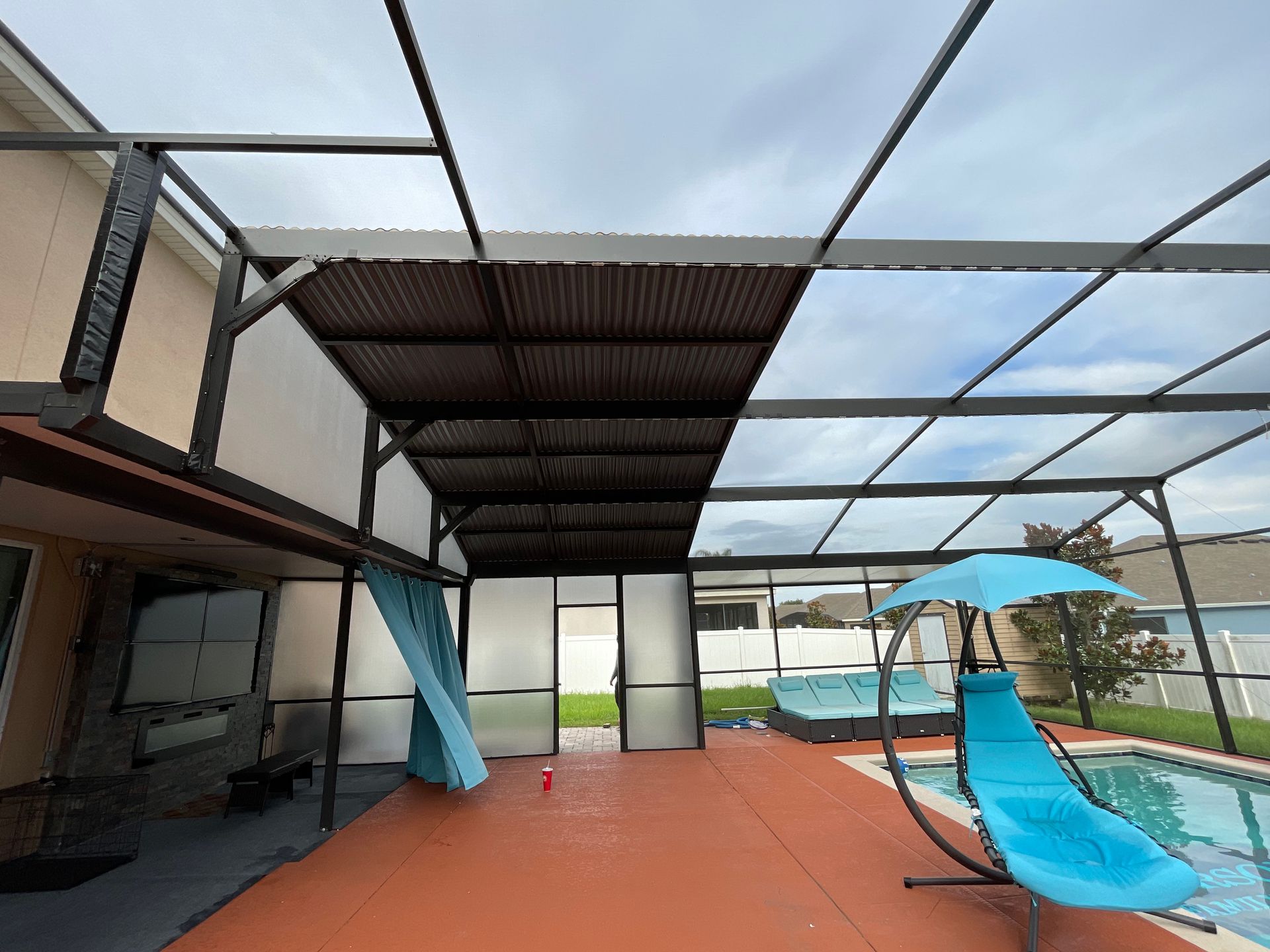 A patio pool enclosure with a partial metal roof, concrete deck, blue lounge chair, and a teal privacy curtain.