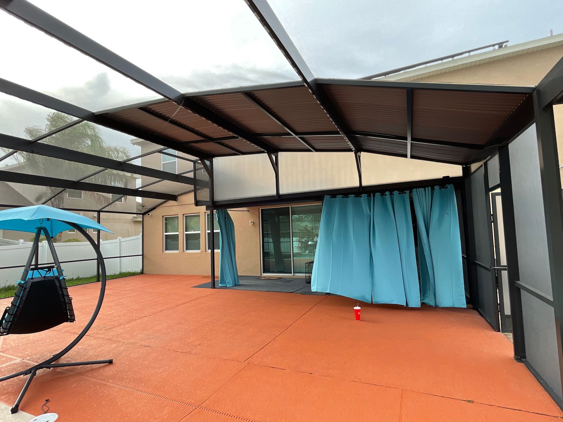 A patio with orange flooring, a black-framed screen enclosure, a blue hanging swing chair, and light blue curtains.