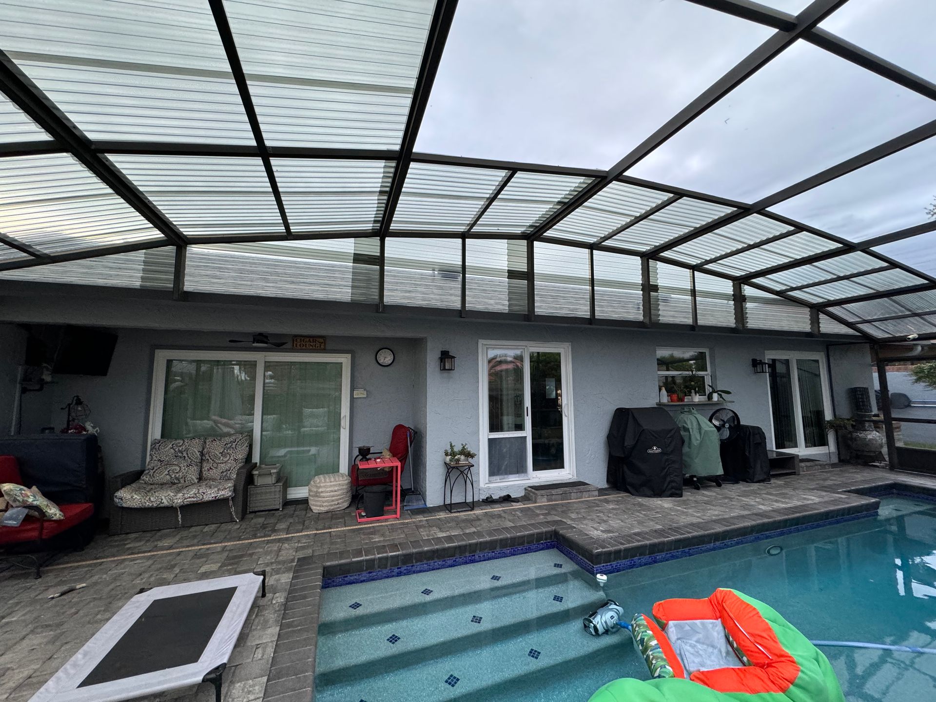 A covered backyard pool patio with gray stucco walls, sliding glass doors, a loveseat, grill, and steps into the pool.