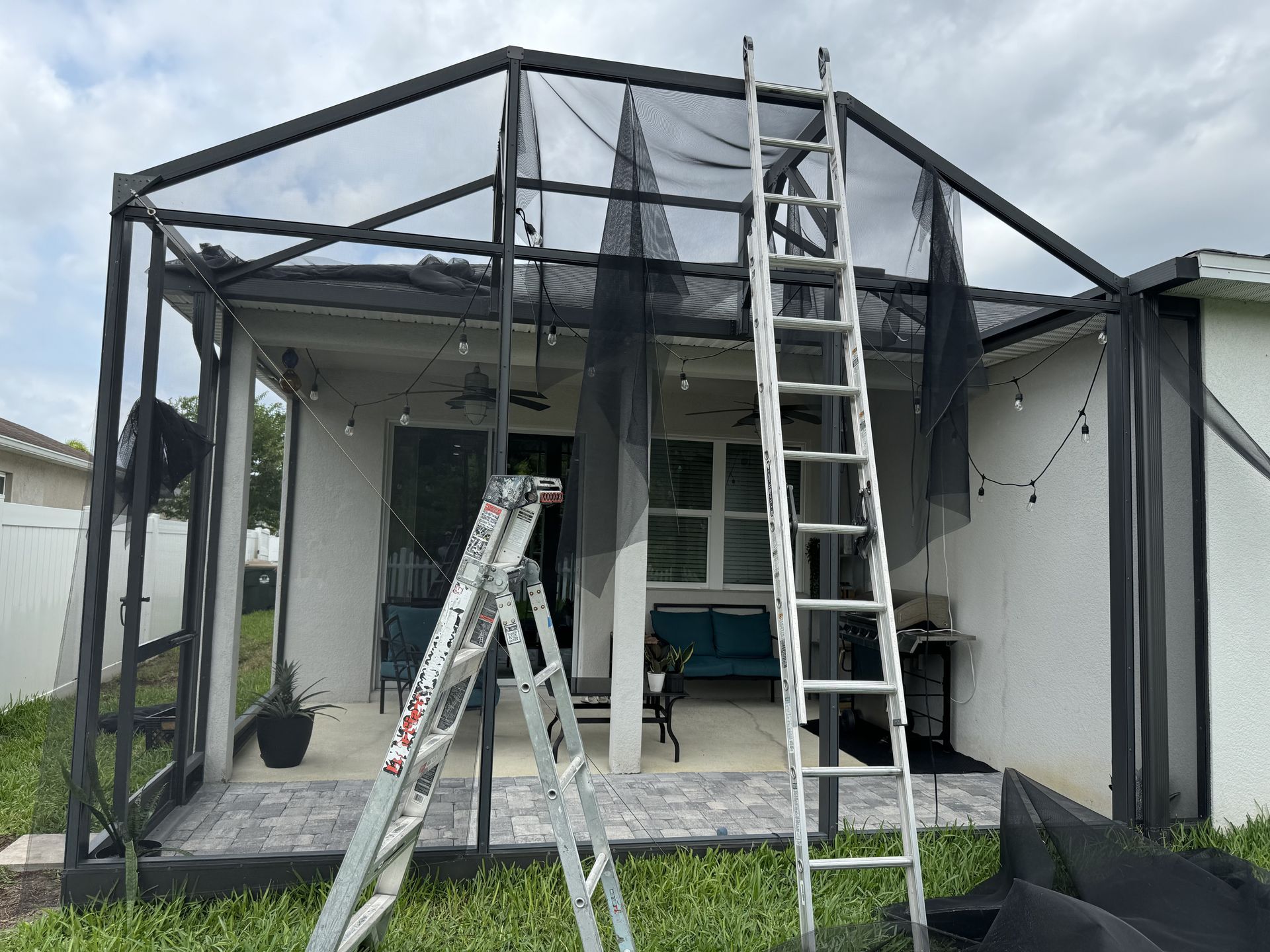 A metal frame patio enclosure with torn screen panels, featuring two ladders positioned against the structure.