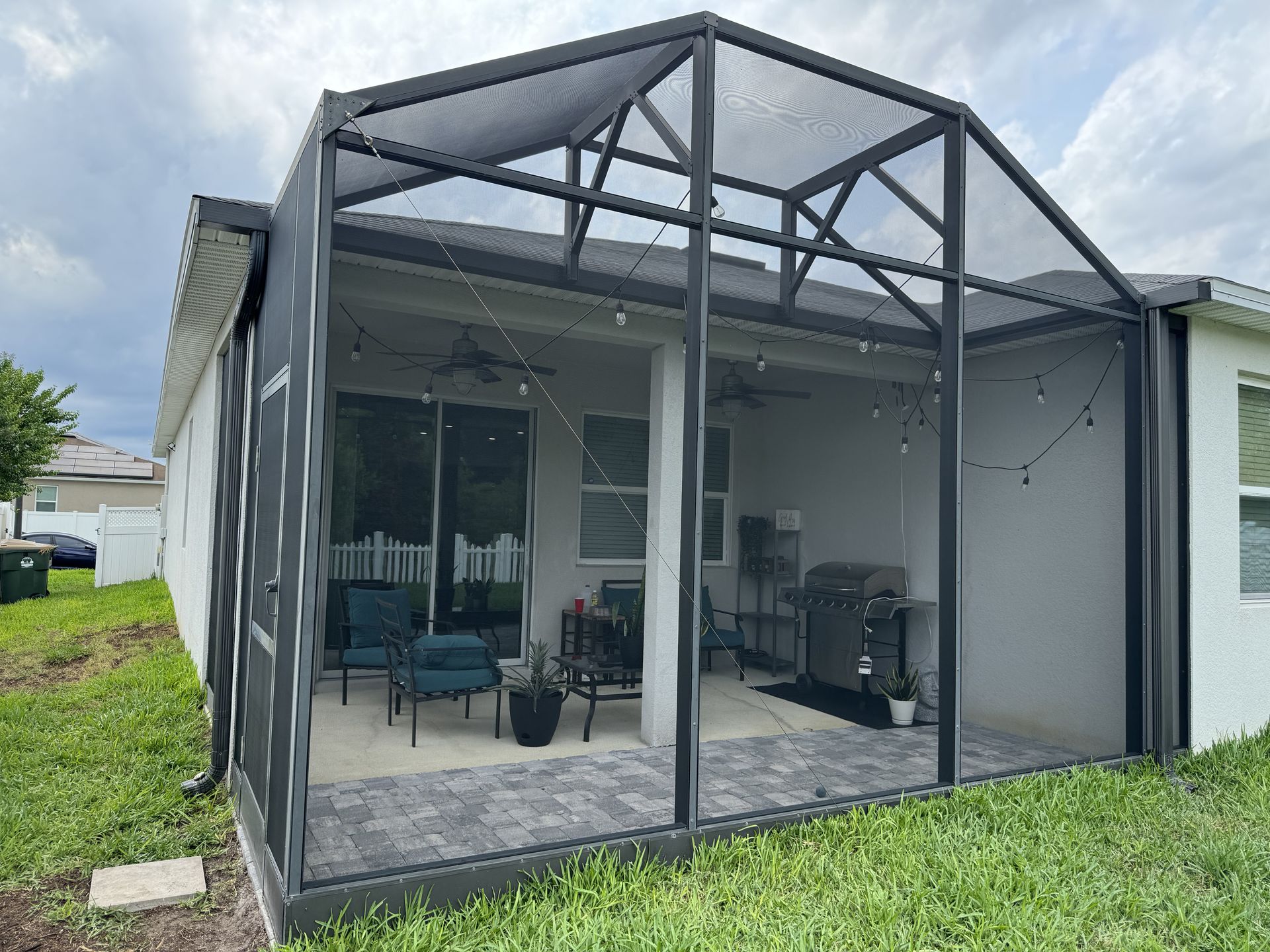A screened-in backyard patio featuring paved floors, outdoor seating, a grill, and string lights under a gabled roof.