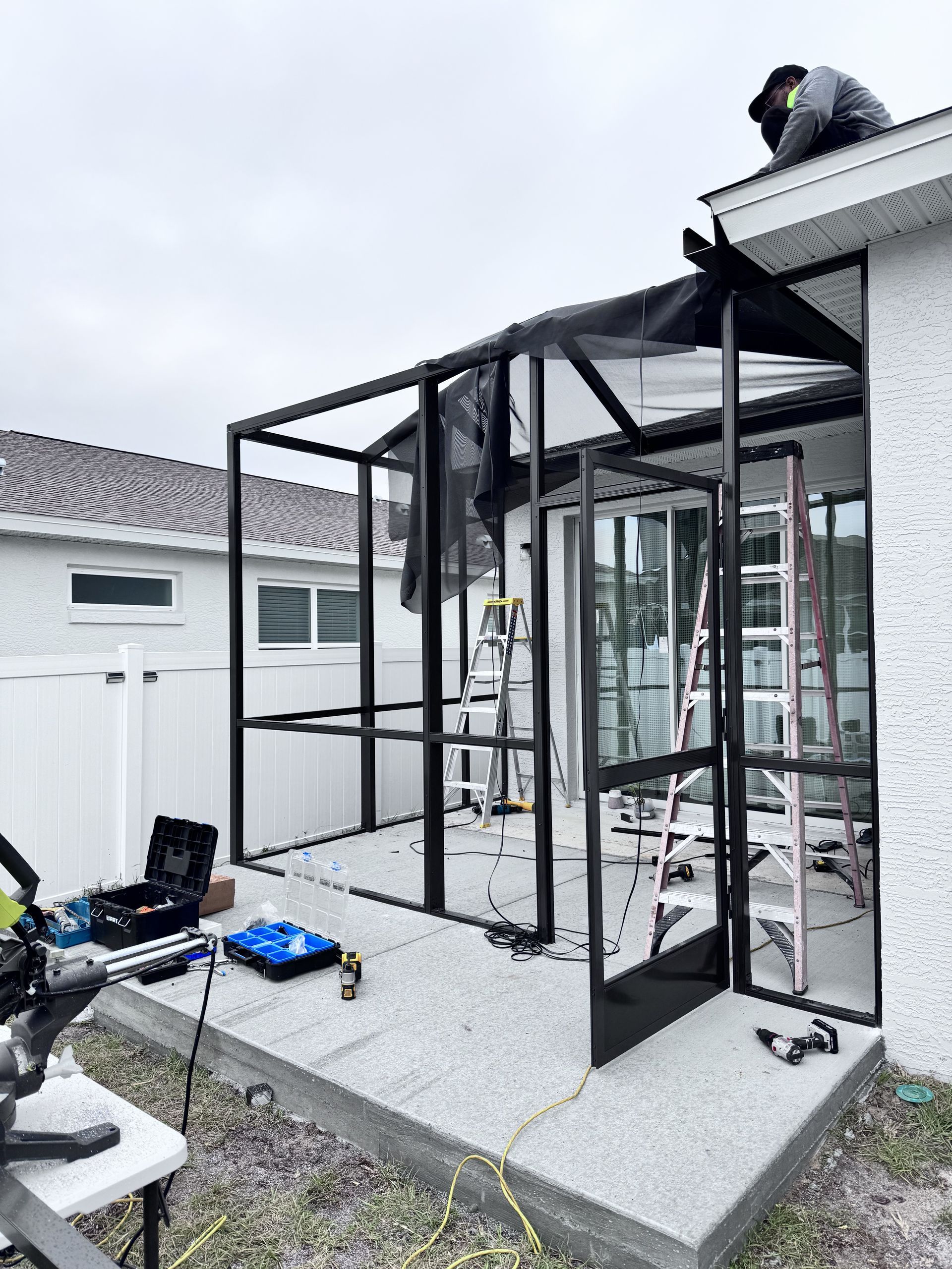 A worker installs a metal screen enclosure frame on a concrete patio at the back of a house.