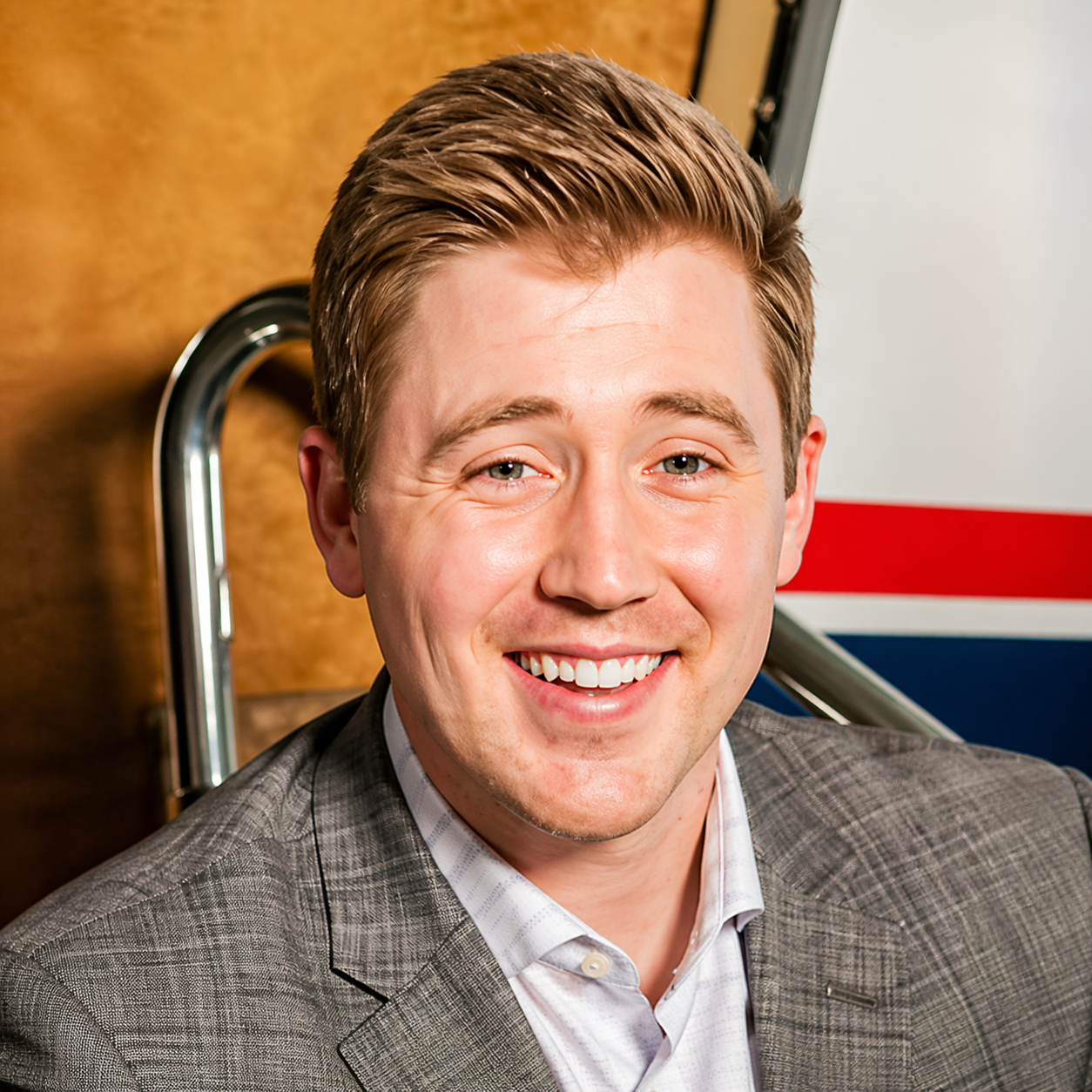 Smiling man in a gray blazer seated indoors beside a railing and red-blue wall stripe