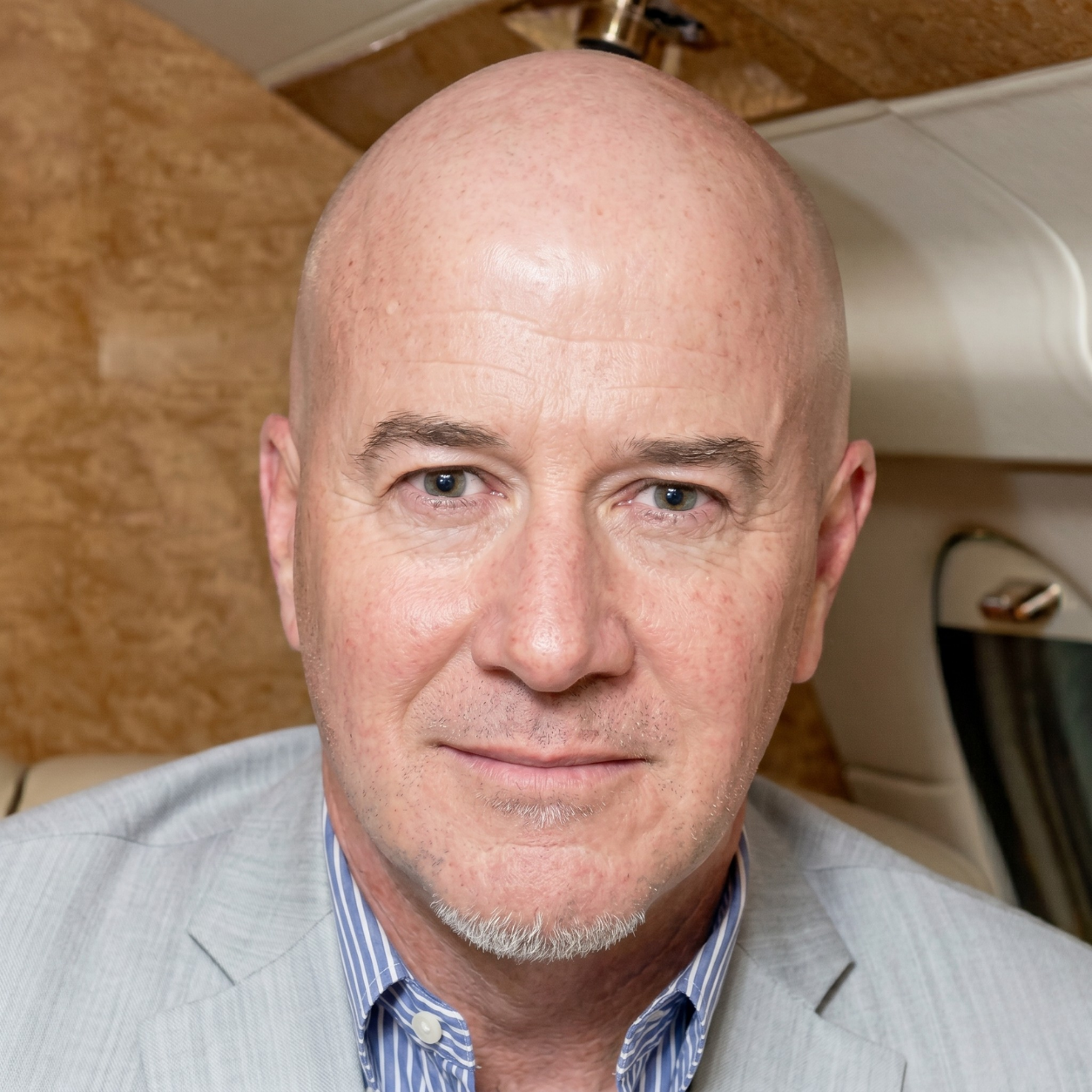 Close-up portrait of a bald man in a light jacket, seated inside a car, looking at the camera
